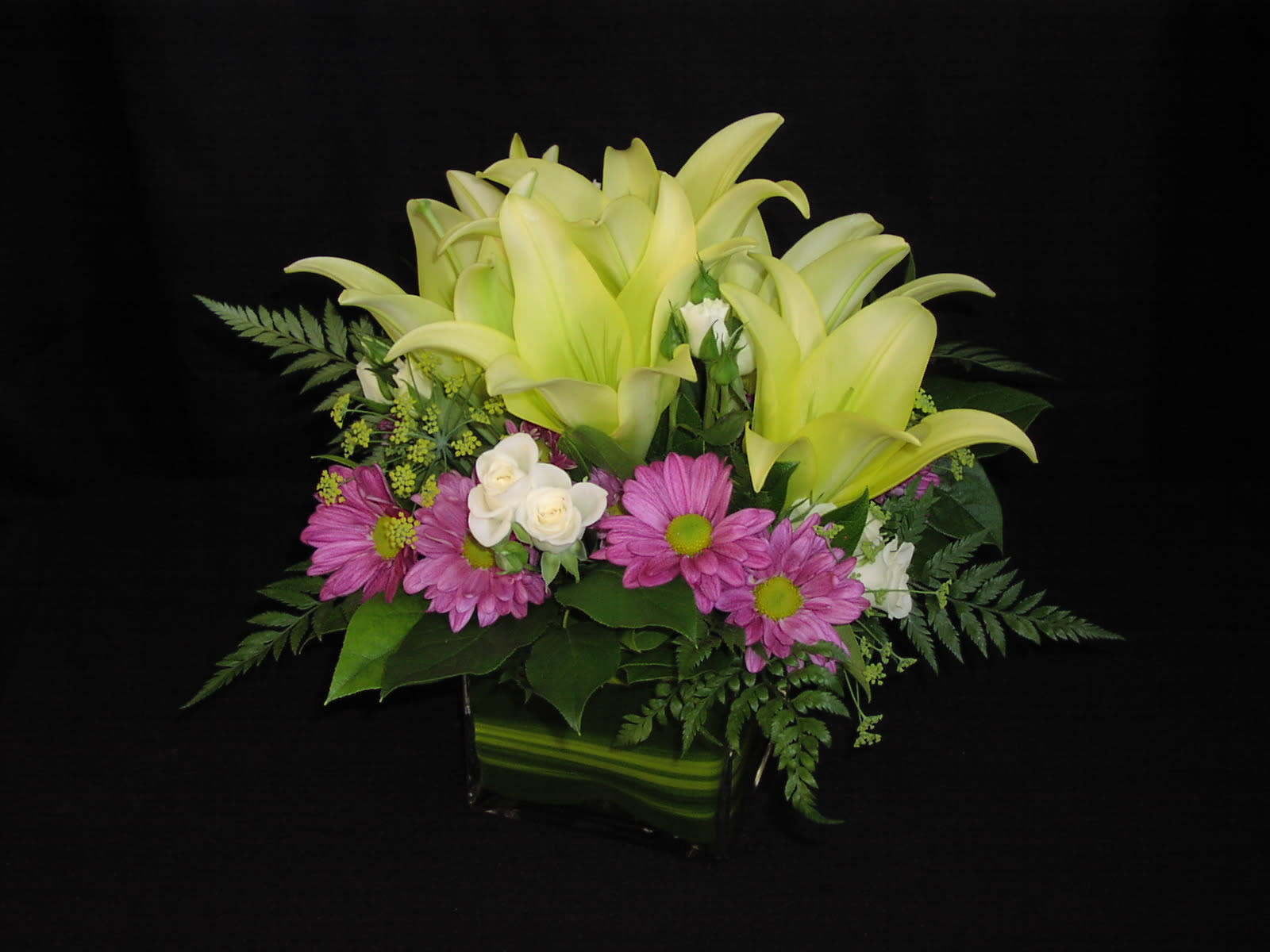 Modern Lily Cube with Spray Roses - A clean design shows off these gorgeous lilies and delicate little spray roses with hints of the season's best filler flowers in a premium clear glass cube vase with a tropical ti leaf wrapped inside.