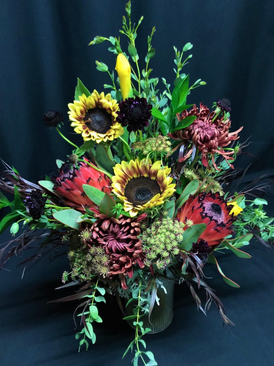 Paprika Protea Pre-Autumn Mix - Rustic Metal Vase of tawny paprika and burgundy flowers, protea, premium Ring of Fire Sunflowers and early autumn favorites.* Subject to availability. Long Lasting! Because every arrangement is made fresh to order, you will always receive a custom one-of-a-kind creation at Flower Fiesta full service florist.  