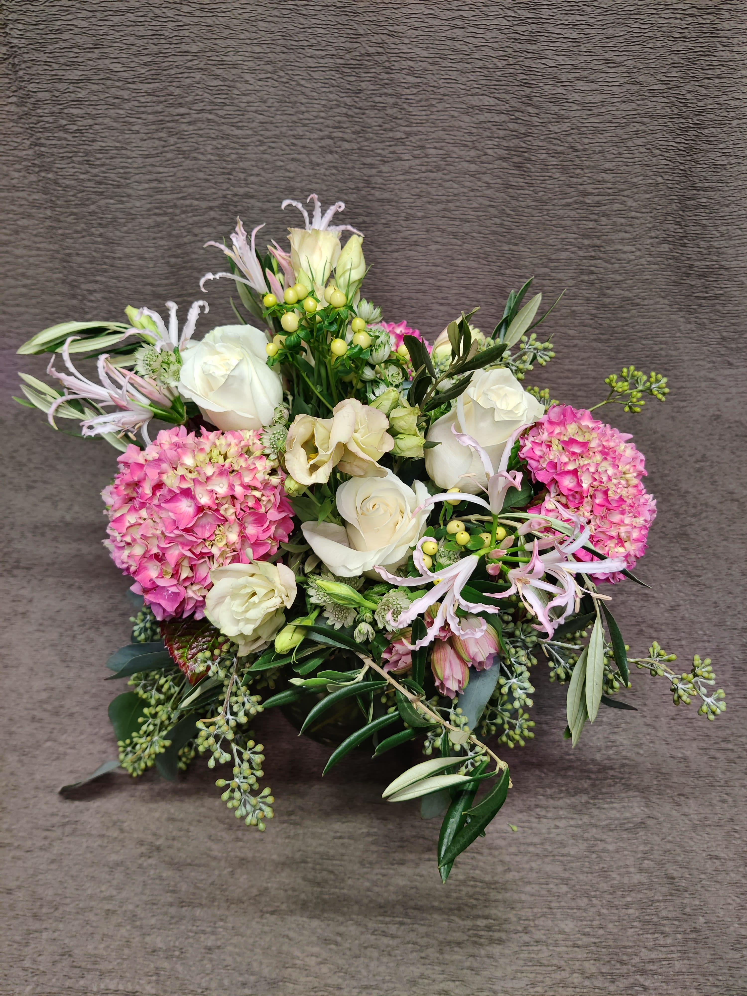 Olive centerpiece pinks and whites  - Flowers all the way around in a centerpiece design for a living room coffee table or dining table for 6.  Medium size bowl of flowers. Premium assortment will vary. Natural artistic freshly gathered look perfect for an anniversary or even a wedding centerpiece.   With a lovely view all around. Grey green olive foliage as seasonally available. Roses hydrangea and/or similar premium flowers in shades of pinks and whites.     