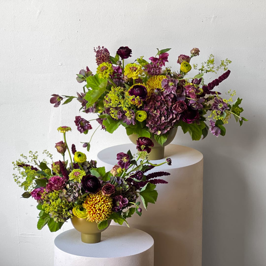 Two purple and yellow floral arrangements in gold vases on white pedestals