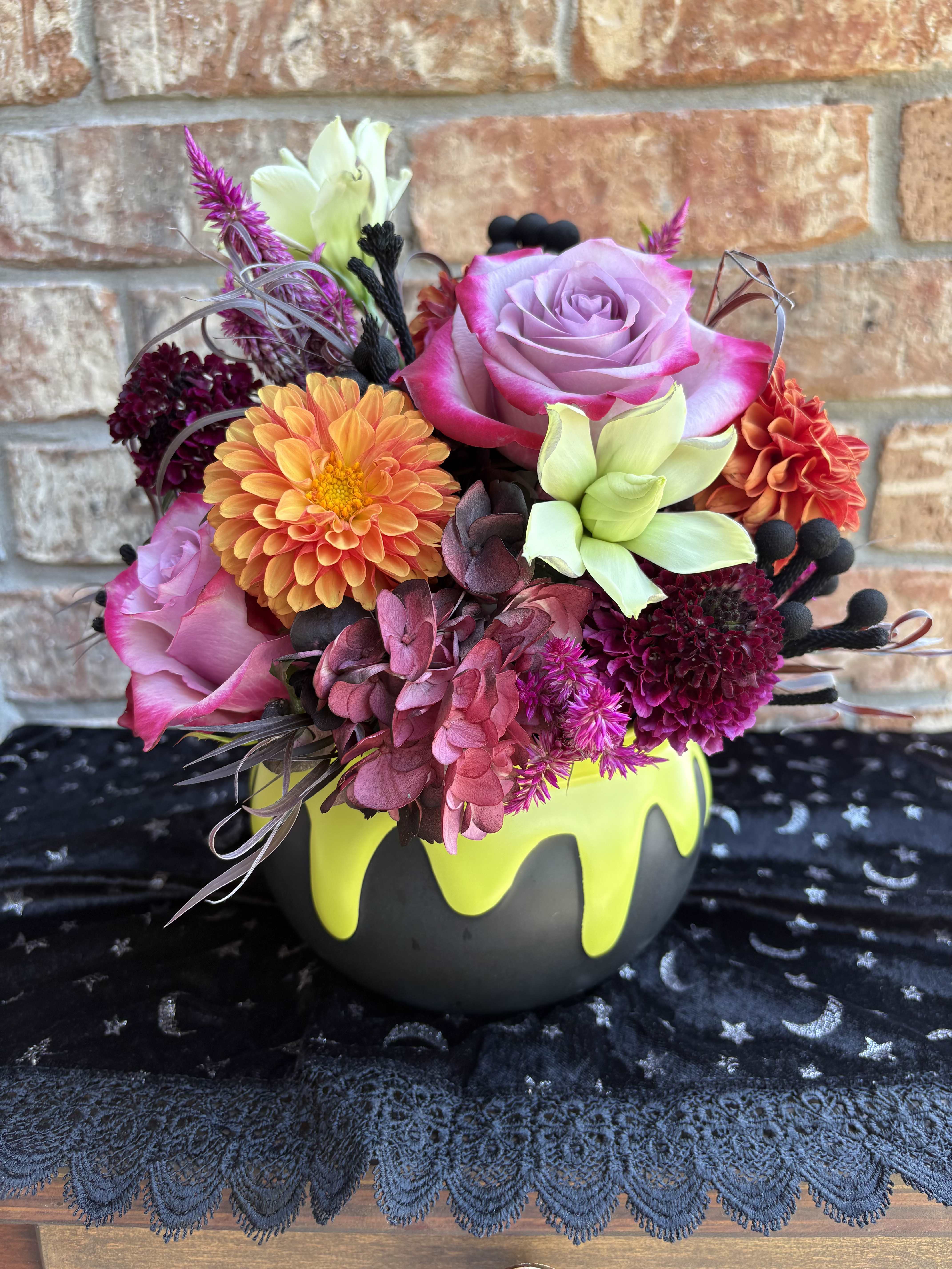 Fall Under My Spell - A whimsical Halloween arrangement designed in a black cauldron-style baking container, brimming with autumn’s most enchanting blooms. orange dahlias, purple roses, velvety lisianthus, and deep purple scabiosa swirl together with pops of green and dramatic black accents. Celosia and brunia add rich texture, while lush hydrangea spills like a potion in bloom. A spellbinding centerpiece that brings a groovy, moody glow to any table or entryway this season.