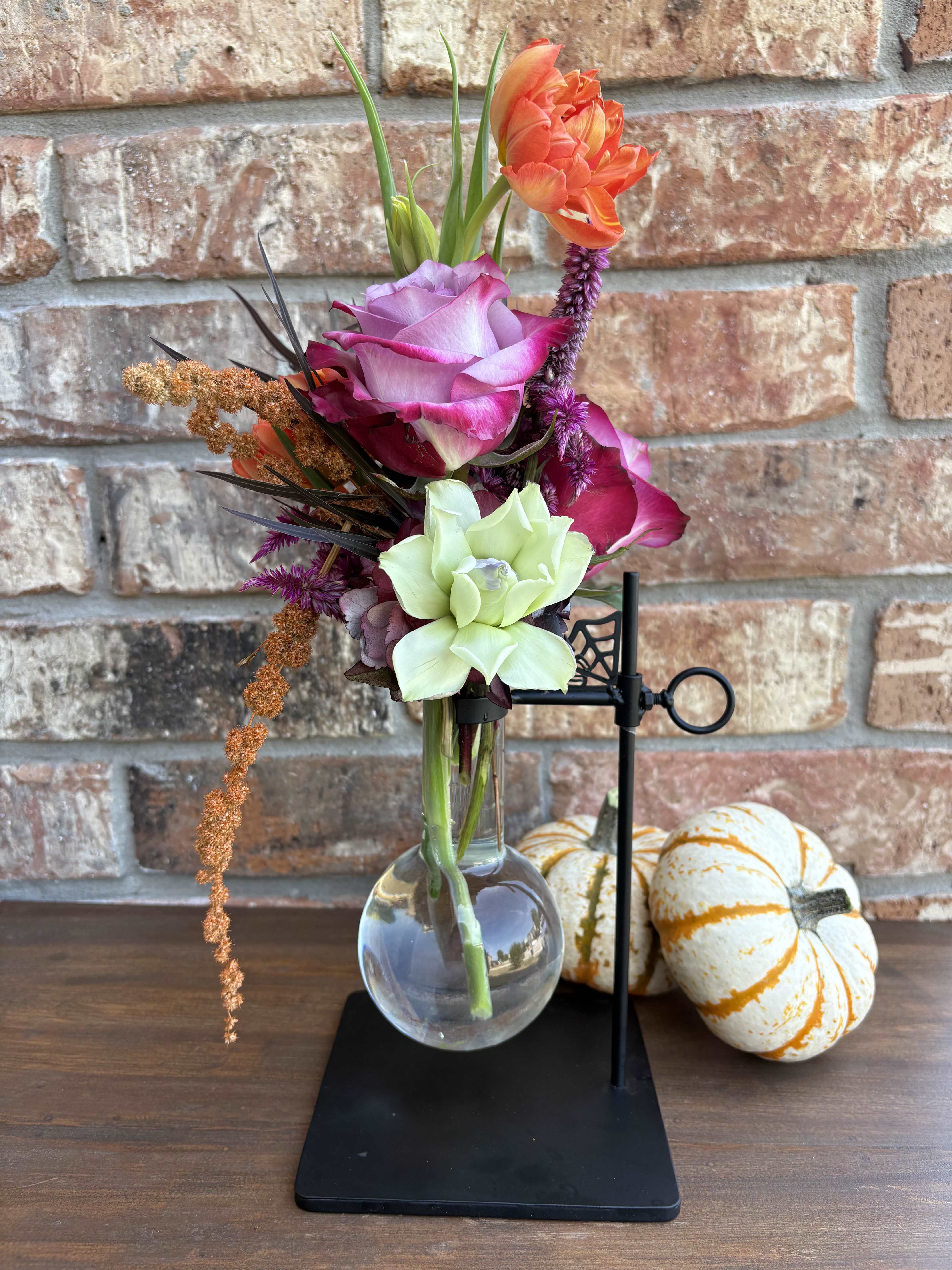 Moonlit Mixture - A bewitching blend of blooms that feels equal parts spell and science. Deep black-tinted hydrangea and burgundy celosia set a dramatic base, while velvety purple roses and lisianthus weave in midnight mystery. Bright orange tulips flicker like candlelight, balanced by subtle touches of fresh green for contrast. Designed in a sleek scientist’s glass container, this arrangement channels retro potion-bottle vibes—perfect for adding a dash of spooky sophistication to your Halloween season.