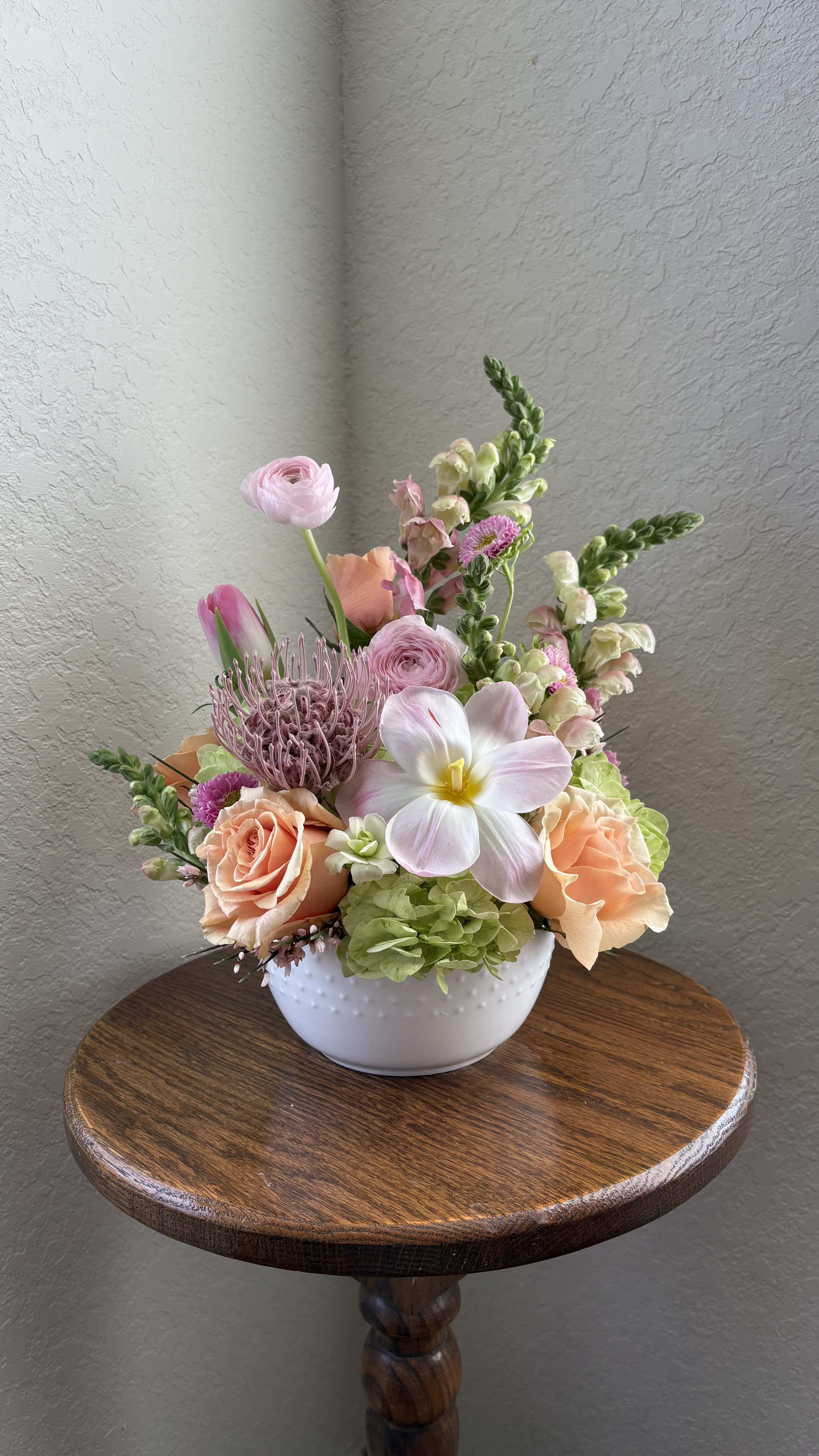 Pastel floral arrangement in a white bowl vase on a wooden table.
