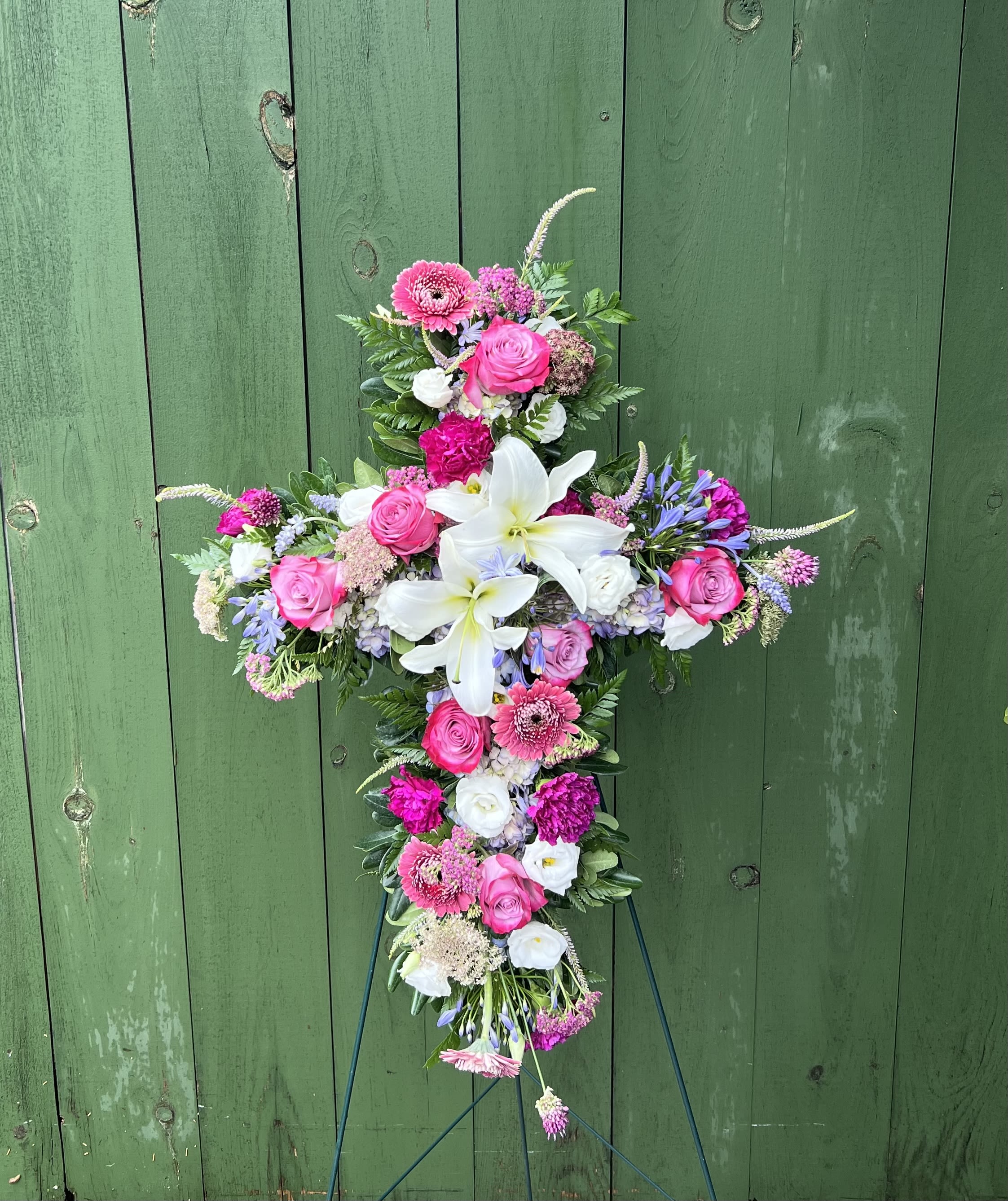 Angelic Garden Cross - Celebrate a life beautifully lived with the Angelic Garden Cross, a graceful floral tribute that reflects faith, love, and everlasting remembrance. This elegant standing cross features fragrant white lilies, rich purple roses, and a delicate blend of soft pink and white blooms nestled among premium seasonal greenery. 