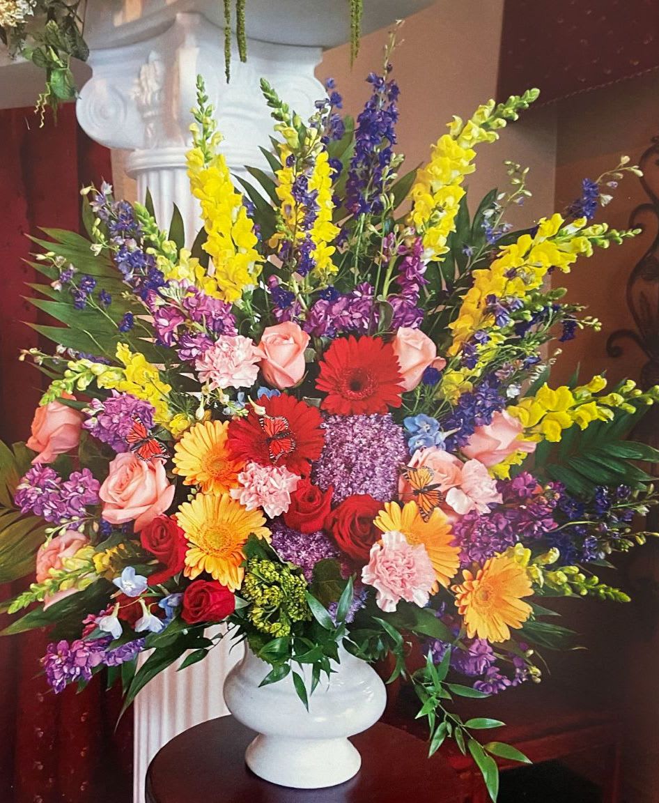 Tall mixed arrangement of bright roses, gerbera daisies, carnations and snapdragons in a white vase.
