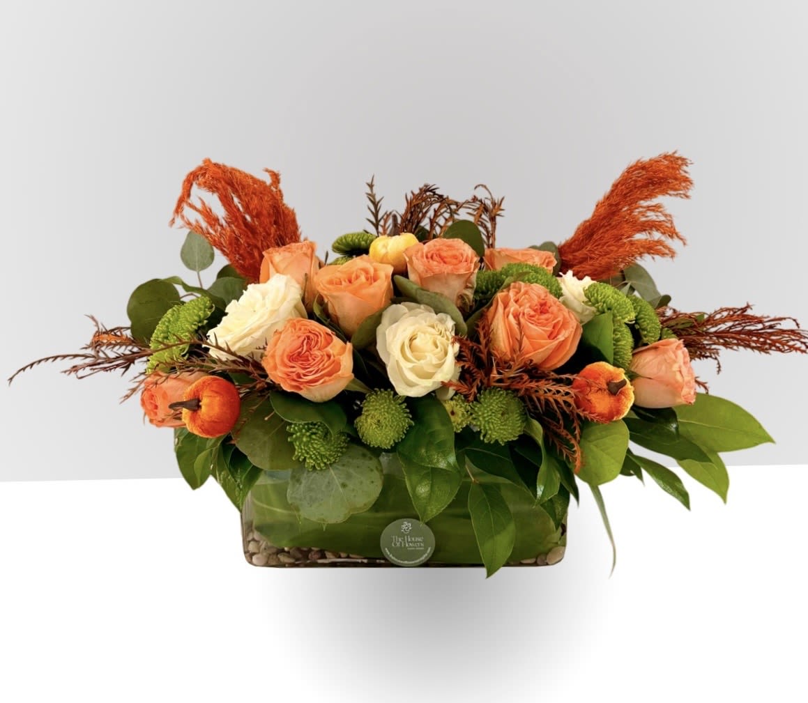 Harvest Glow Centerpiece by The House of Flowers - Celebrate Thanksgiving with our Harvest Glow Centerpiece: featuring peach and cream roses, lush greenery, and warm autumn textures. Designed in a modern glass base (5" x 5"), this arrangement brings elegance, warmth, and seasonal charm to your holiday table.”