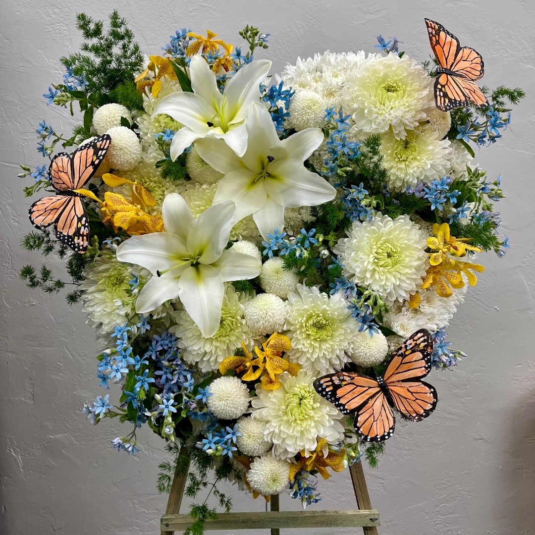 Heart-shaped floral arrangement with white lilies, chrysanthemums, blue flowers, and butterfly accents