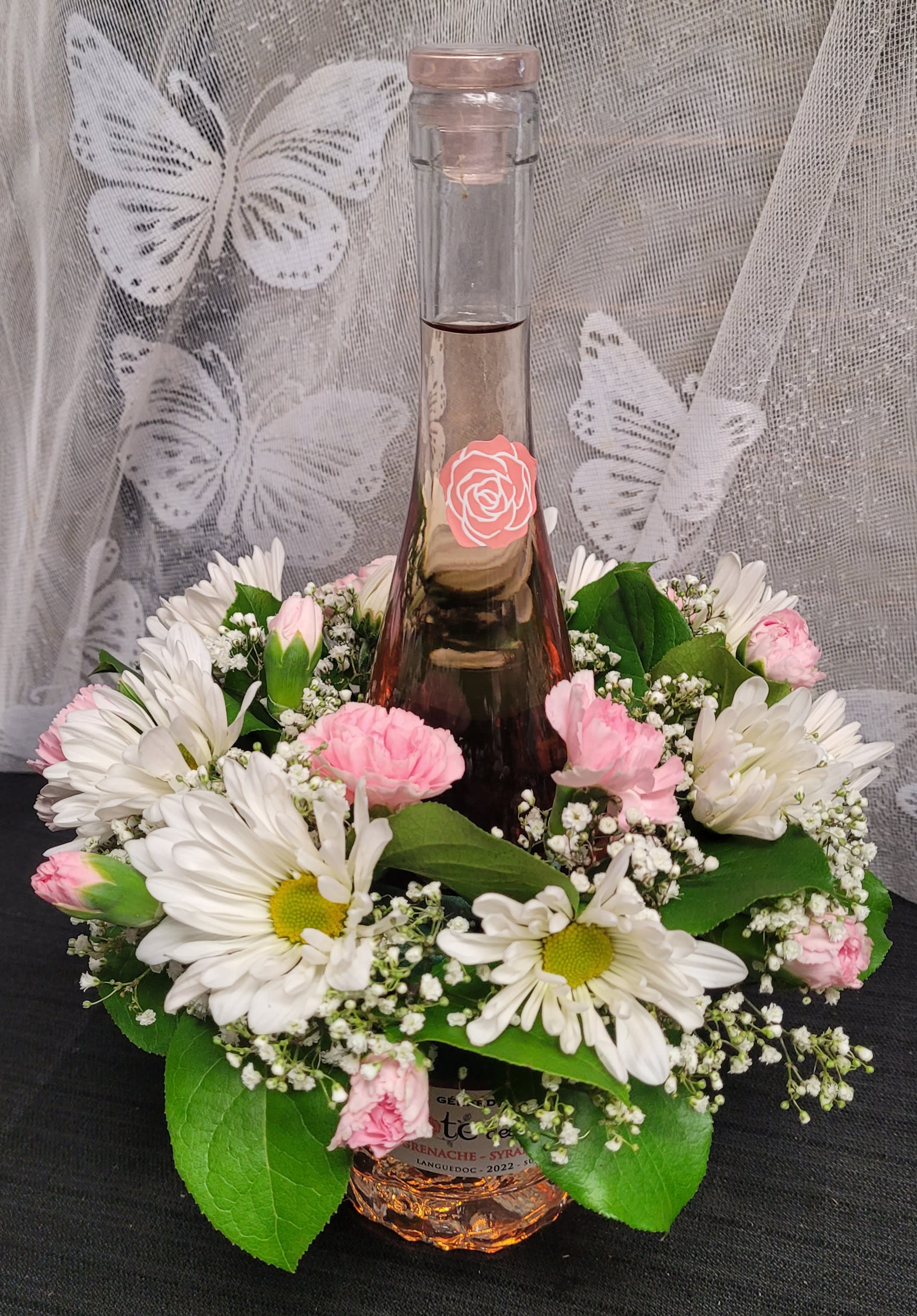 Whimsy wine bottle - A tall glass bottle of rosé rises from the center of a low, rounded floral collar-a playful, celebratory flower arrangement perfect as a Mother's Day gift, anniversary surprise, or holiday hostess present. The vertical bottle and compact ring combine the look of a bouquet with the charm of a gift, making it an ideal way to say "thank you," celebrate love and romance, or toast New Year moments with someone special.  The wreath-like collar mixes bright magenta and hot pink carnations, soft pink roses, magenta chrysanthemums, white daisy-style blooms with sunny yellow centers, pale alstroemeria, and delicate white filler flowers set against fresh green leaves for a lush, textured look. The arrangement sits around a tall clear glass wine bottle with a stopper and rose emblem; when removed, the floral ring can double as a centerpiece or hold a candle for a cozy table accent. Must be 21 to purchase; unless you request otherwise, a rosé will be selected.