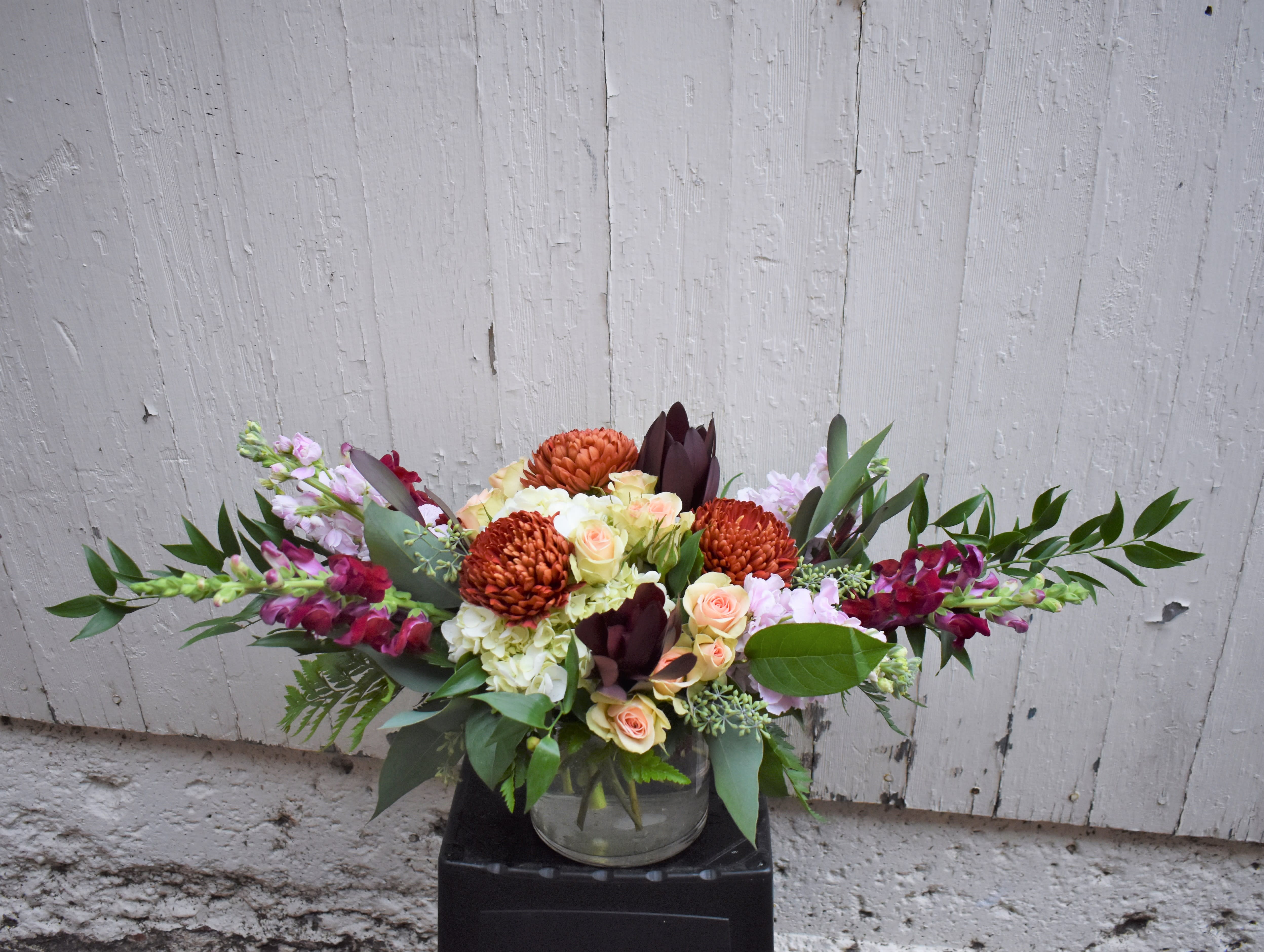 Wild Wanderings - Luxurious greenery gracefully arcs and pulls the eye towards a mix of blush spray roses, bronze mums, burgundy Safari Sunset, white hydrangea pink stock, and wine snapdragons. This is a wonderful bouquet for elegantly celebrating any autumn occasion! 