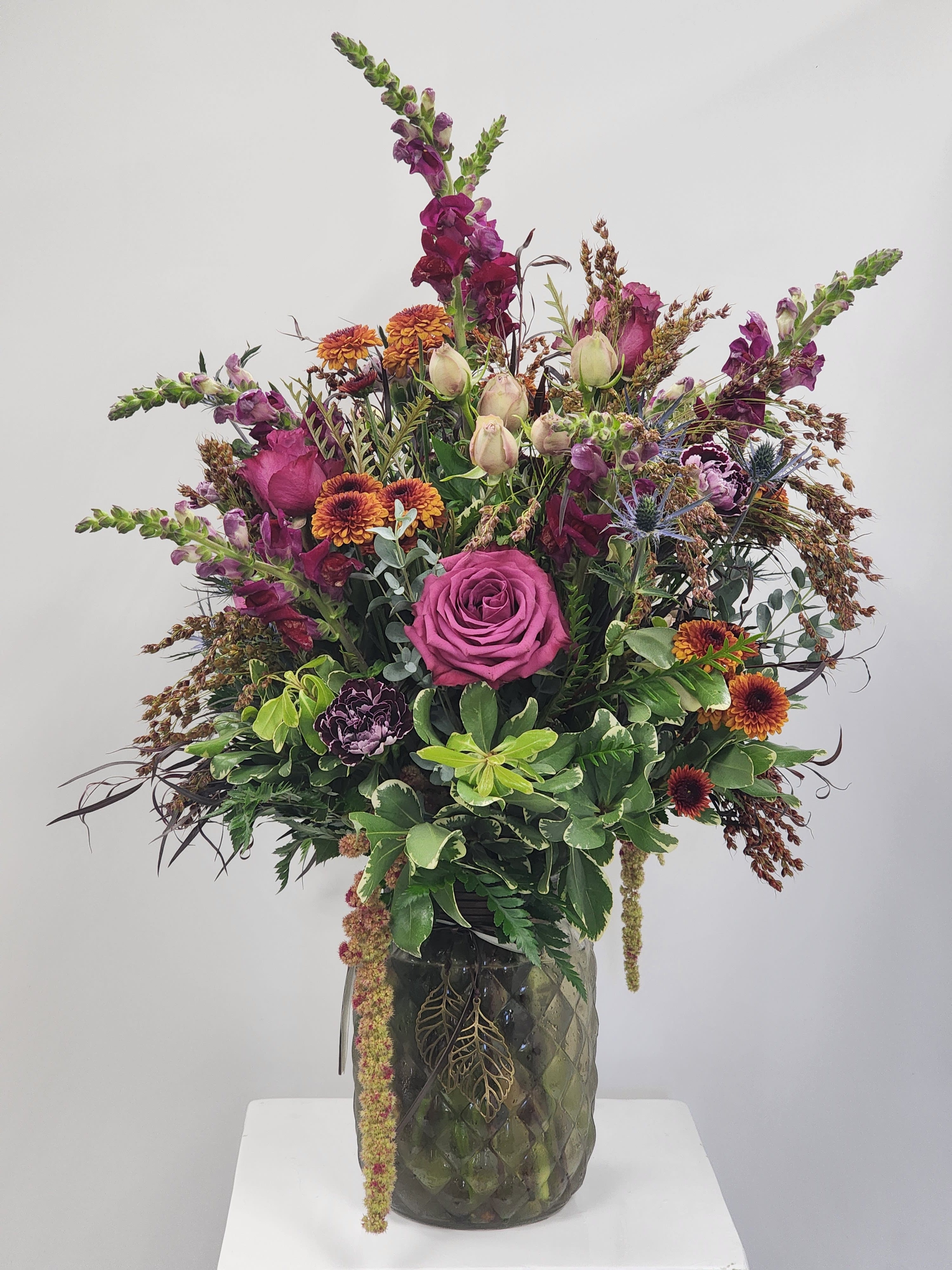 Autumn Orchard - Select this single-sided autumnal arrangement. Includes snapdragons, roses, pompoms, spray roses, carnations, amaranthus, broomcorn, and thistle in a cute fall vase. Approximately 24" tall by 14" wide.