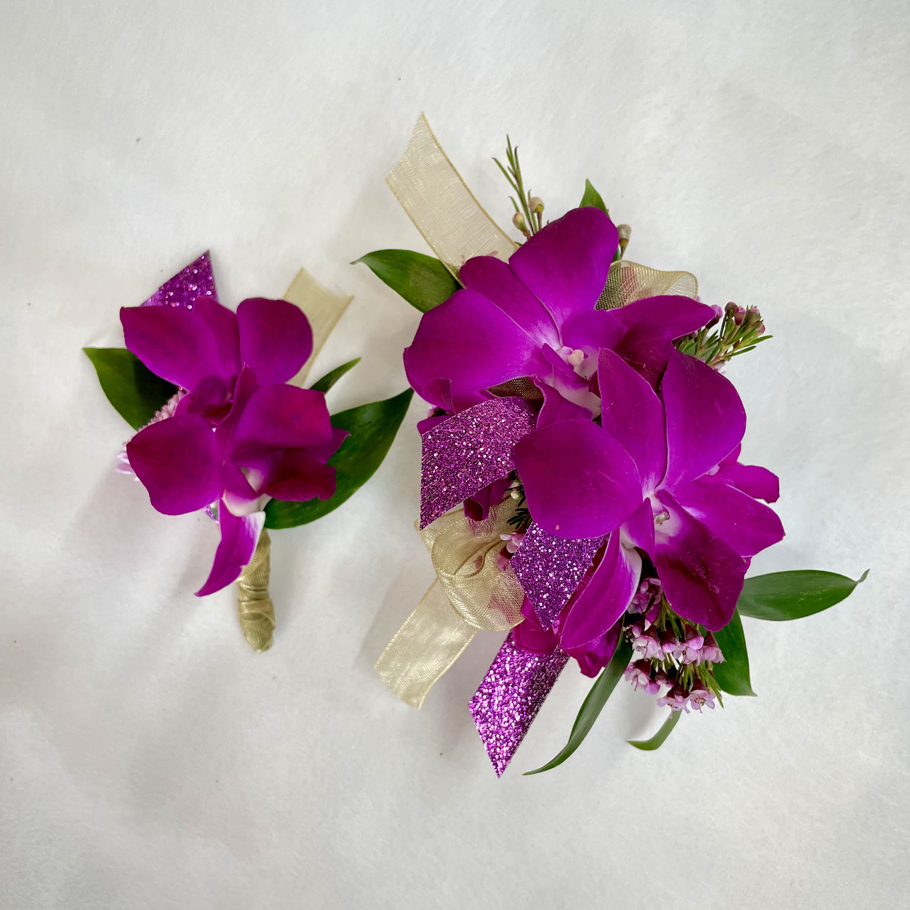 Fuchsia orchid corsage and matching boutonniere with gold ribbon and purple glitter accents on a white background