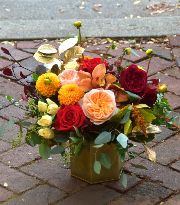 Autumn-tone arrangement of red roses, peach blooms and yellow flowers in a gold cube vase