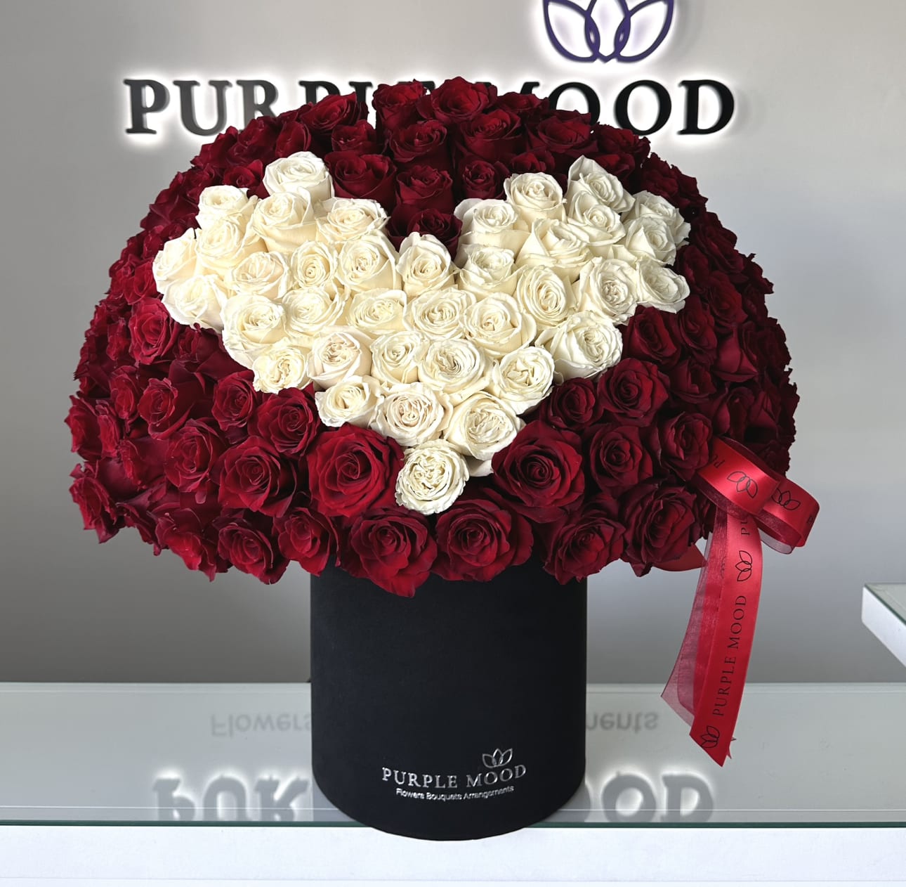 Love Box - Approximately 110-120 roses. Container size - D10.5'x H12.25'  The Love Box is the ultimate statement of passion and devotion. Featuring a grand arrangement of red roses with a heart of creamy white blooms at the center, this design speaks louder than words. Presented in a sleek black box, it’s the perfect gift for anniversaries, Valentine’s Day, or unforgettable romantic gestures.  Key Features:     •    Striking design with a heart motif crafted from roses     •    Presented in a luxury black hat box for a bold, modern look     •    Finished with signature ribbon for an elegant detail     •    A breathtaking gift for anniversaries, proposals, or grand romantic surprises