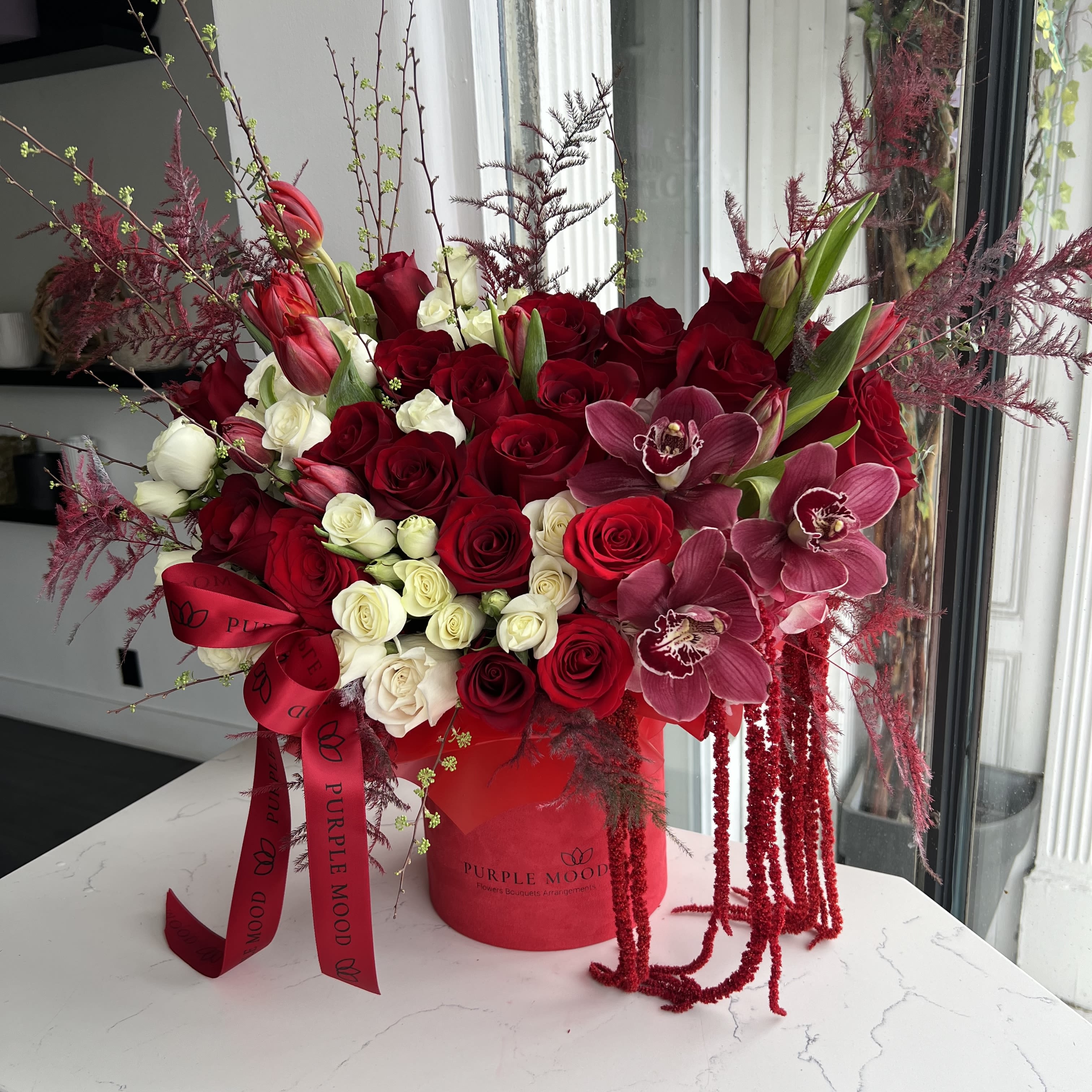 Large red and white rose arrangement with mauve orchids in a red hatbox with cascading red accents.