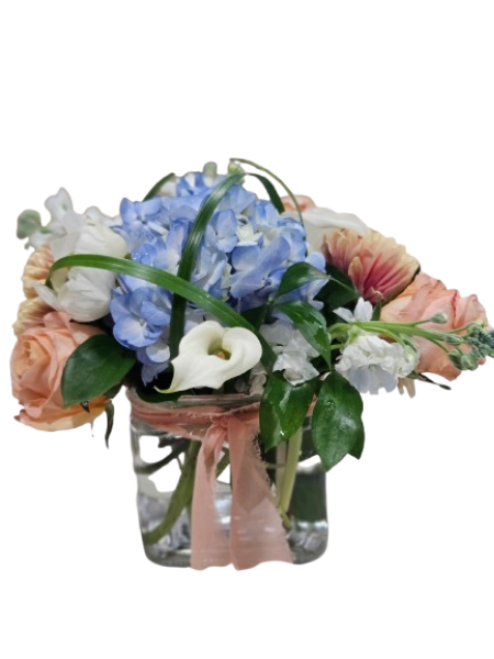 Coastal Whisper Bouquet - Soft, serene, and effortlessly elegant, the Coastal Whisper Bouquet captures the beauty of a breezy seaside day. Featuring lush blue hydrangea at the center, this arrangement is accented with creamy white calla lilies, delicate white blooms, and romantic blush-toned roses for a perfectly balanced, airy look.  Designed in a clear glass vase and finished with a soft blush ribbon, this bouquet brings a calming, coastal charm to any space. It’s a beautiful choice for birthdays, thank yous, sympathy, or simply brightening someone’s day.
