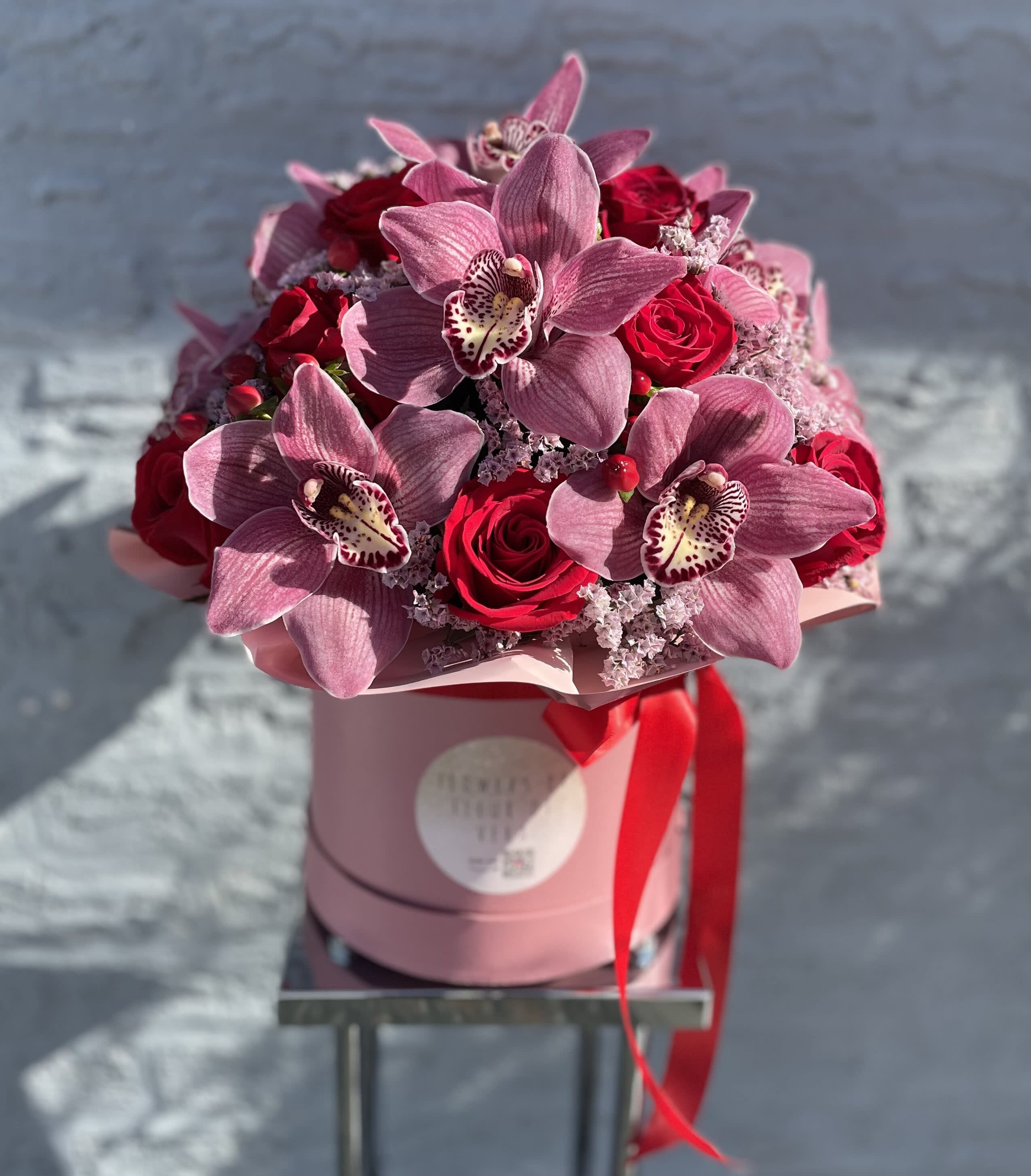 Pink orchids and red roses arranged in a round hatbox with a red ribbon