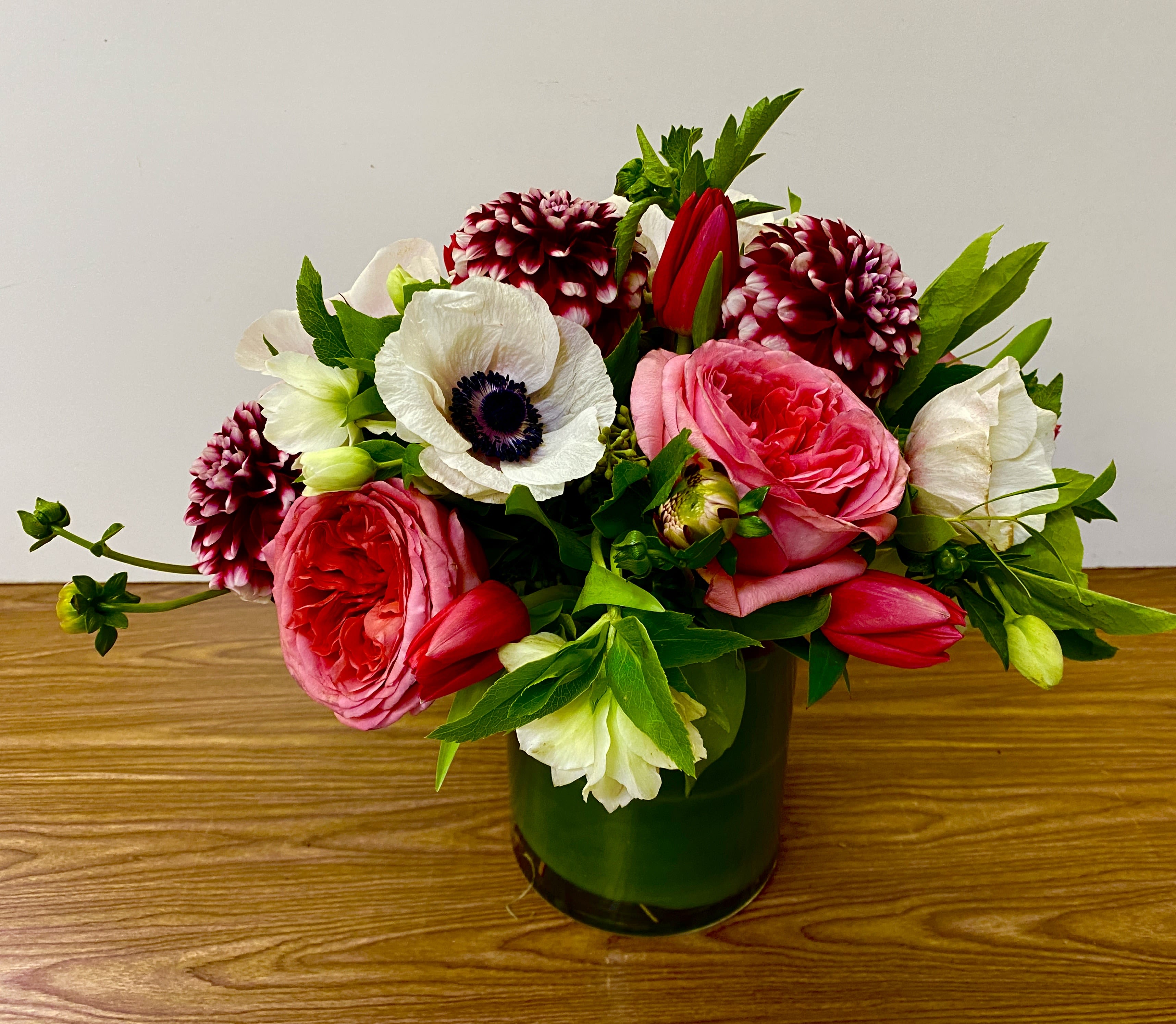 Low pink and white flower arrangement with roses, anemones, tulips, and dahlias in a green glass cylinder vase