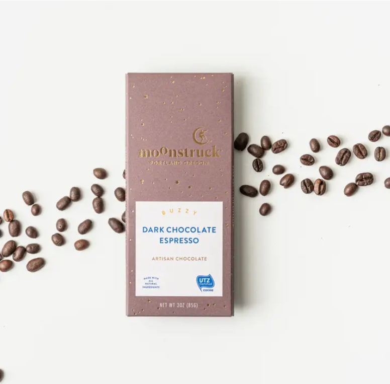 Dark chocolate espresso bar in mauve wrapper with coffee beans scattered on a white surface