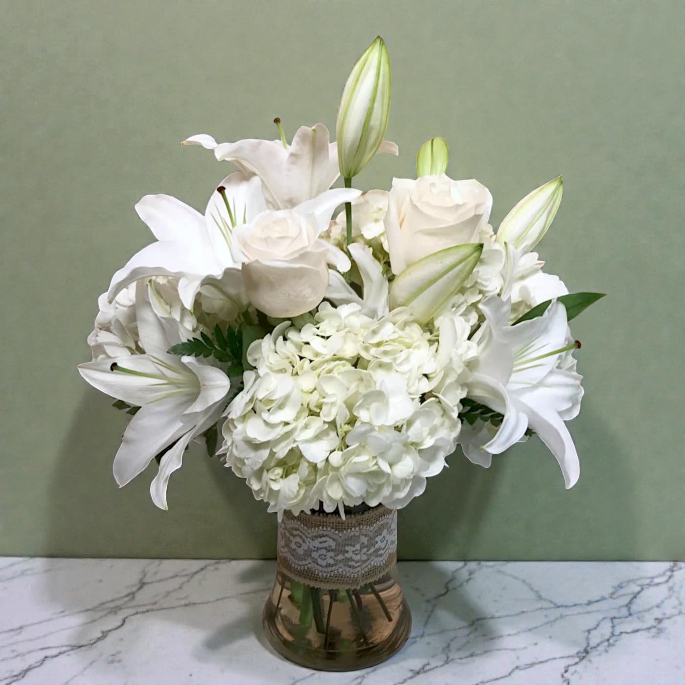 White lilies and hydrangeas arranged in a glass vase with lace trim