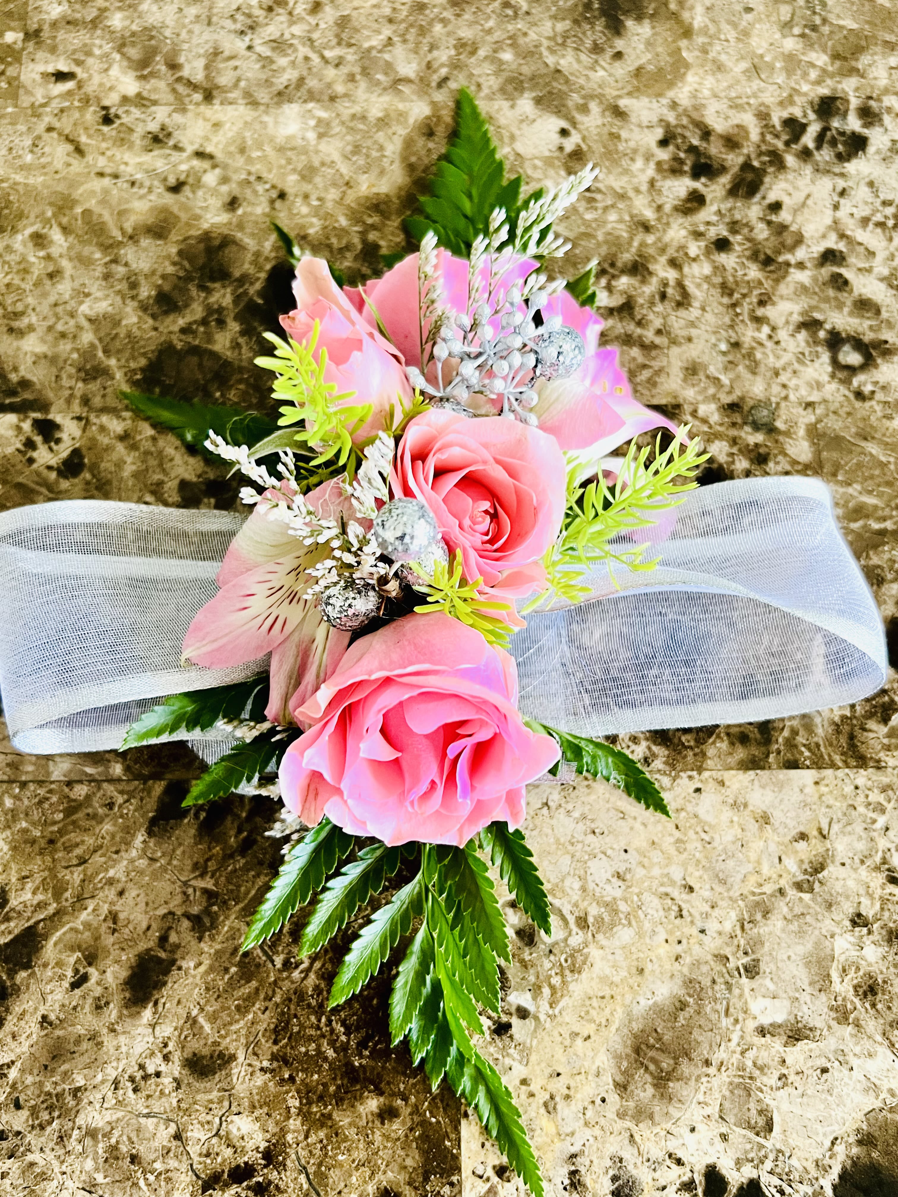 Flower corsage -  Four soft pink roses paired with a pale pink alstroemeria create a quietly romantic wedding corsage designed for brides and attendants. The compact floral cluster sits against bright green foliage and tiny silver decorative berries, with a wide white ribbon trailing behind for a polished, formal finish-perfect for a ceremony, rehearsal dinner, or bridal shower.  