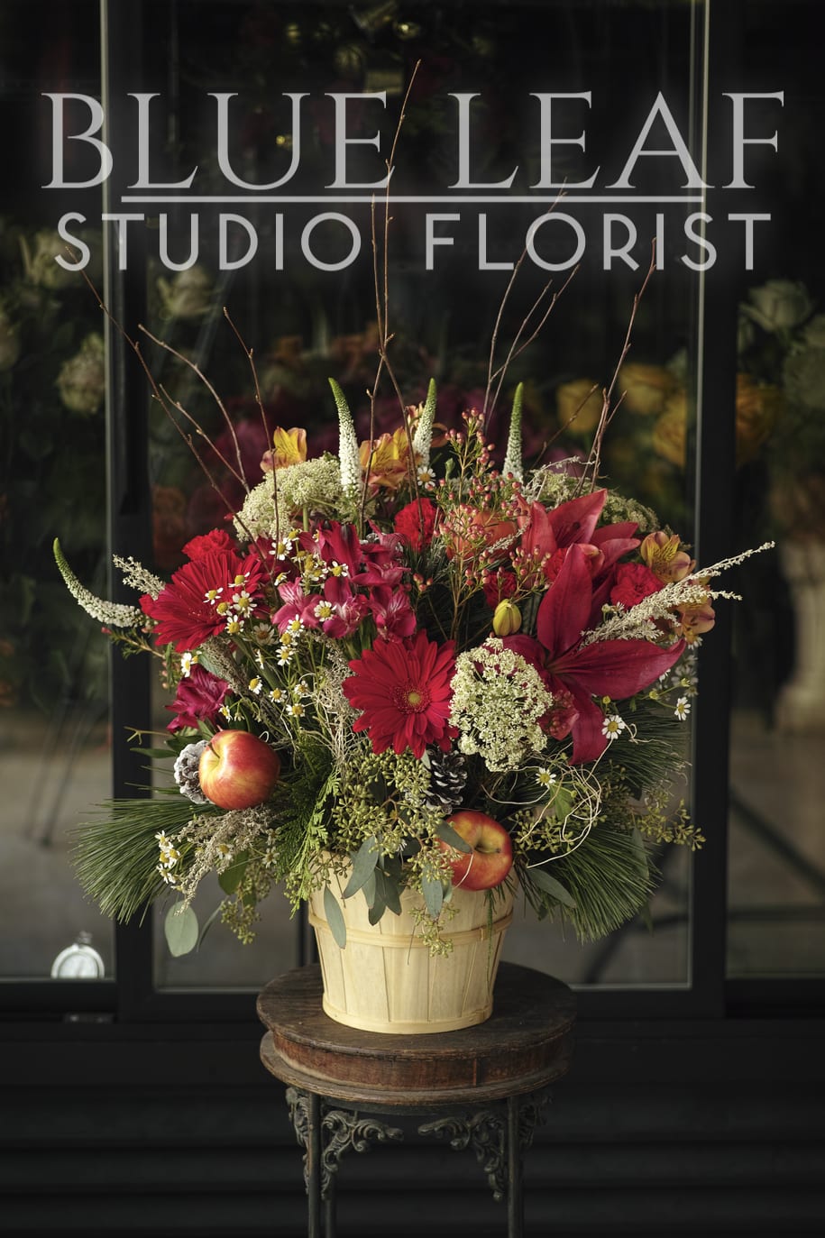 Gala Garden - Natural, country style design in a basket. Arranged with flowers in warm tones, including red gerberas, alstromeria, pops of white, and a fun addition of Gala apples for an autumn feel.