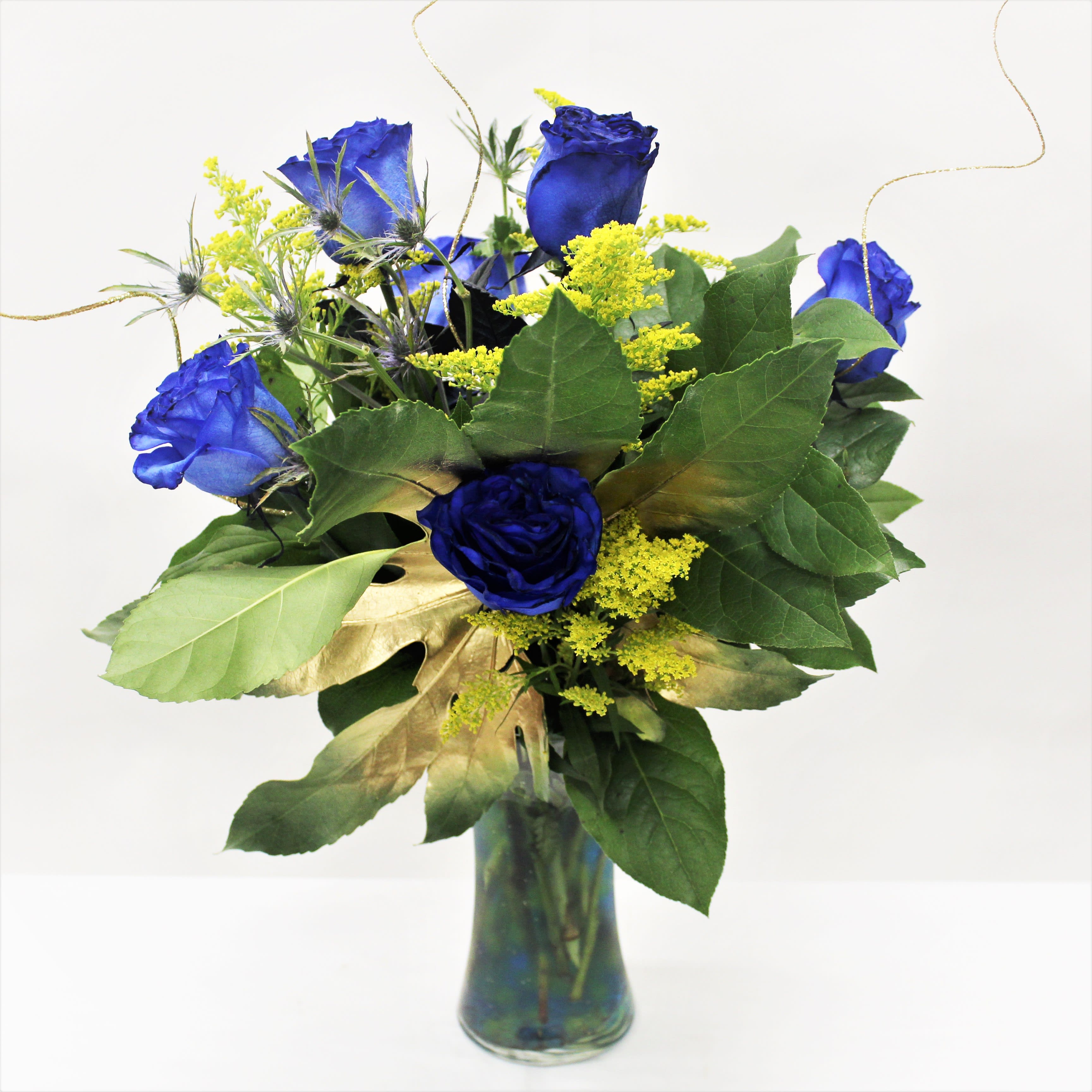 Vase arrangement of vivid blue roses with yellow accents and gold leaves in a clear glass vase.