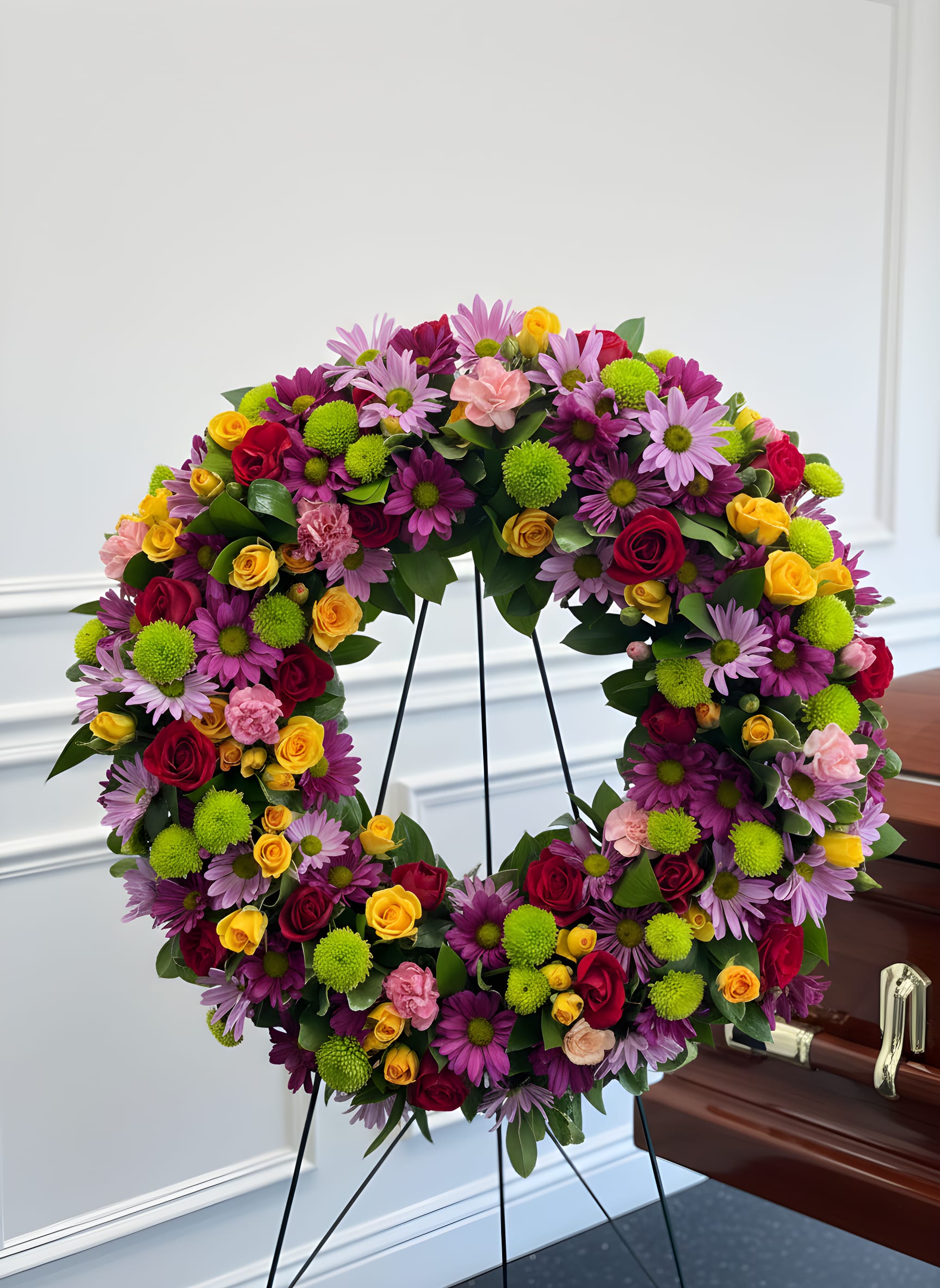Large floral wreath with roses and daisies on a stand beside a casket