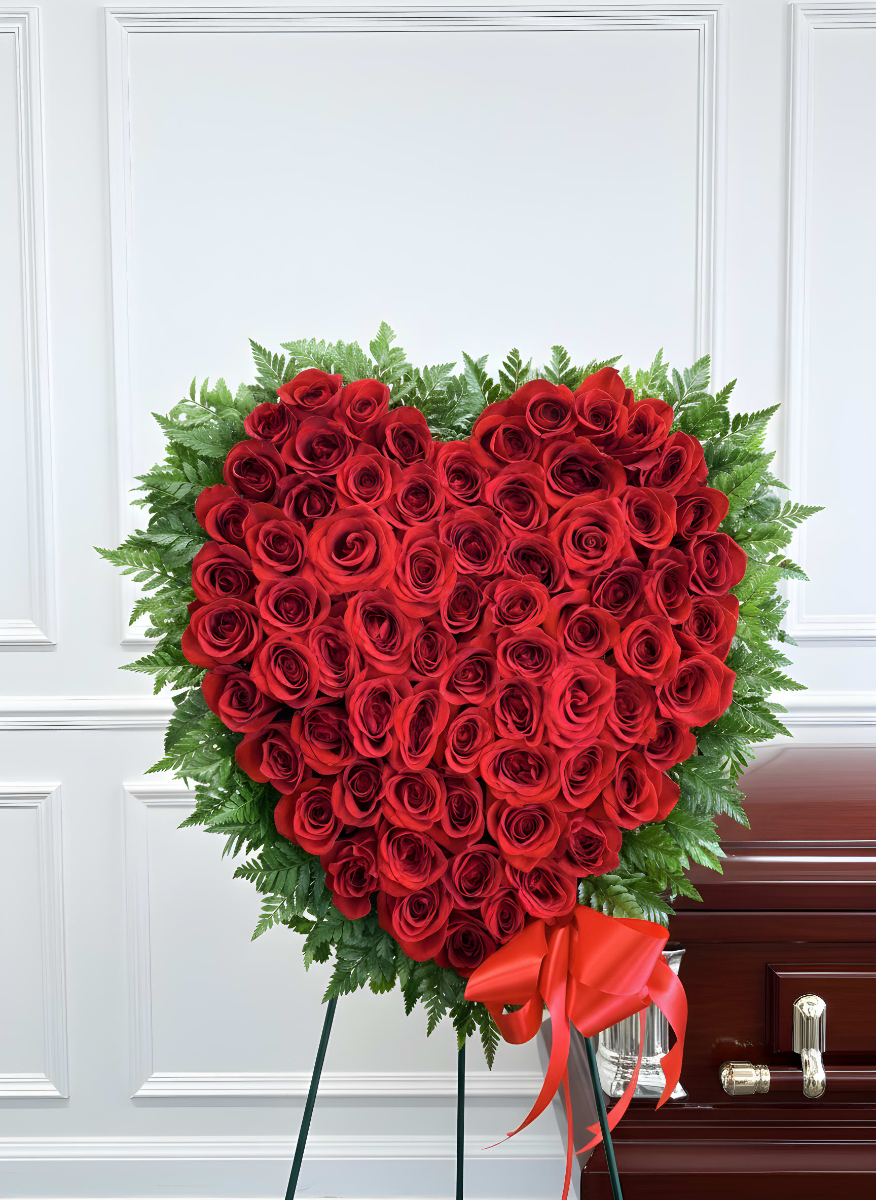 Forever in My Heart Red Rose Standing Spray - Celebrate a life filled with love through our Forever in My Heart Red Rose Standing Spray. This breathtaking heart-shaped funeral tribute is handcrafted with dozens of premium long-stem red roses, symbolizing everlasting love, admiration, courage, and cherished memories. Framed with lush fresh greenery and accented with a luxurious satin red bow, this elegant standing spray creates a powerful expression of love and remembrance.  Designed to stand beautifully beside the casket or at a memorial service, this all-red rose heart is a timeless sympathy arrangement that offers comfort to grieving families while honoring someone who will forever remain in your heart.