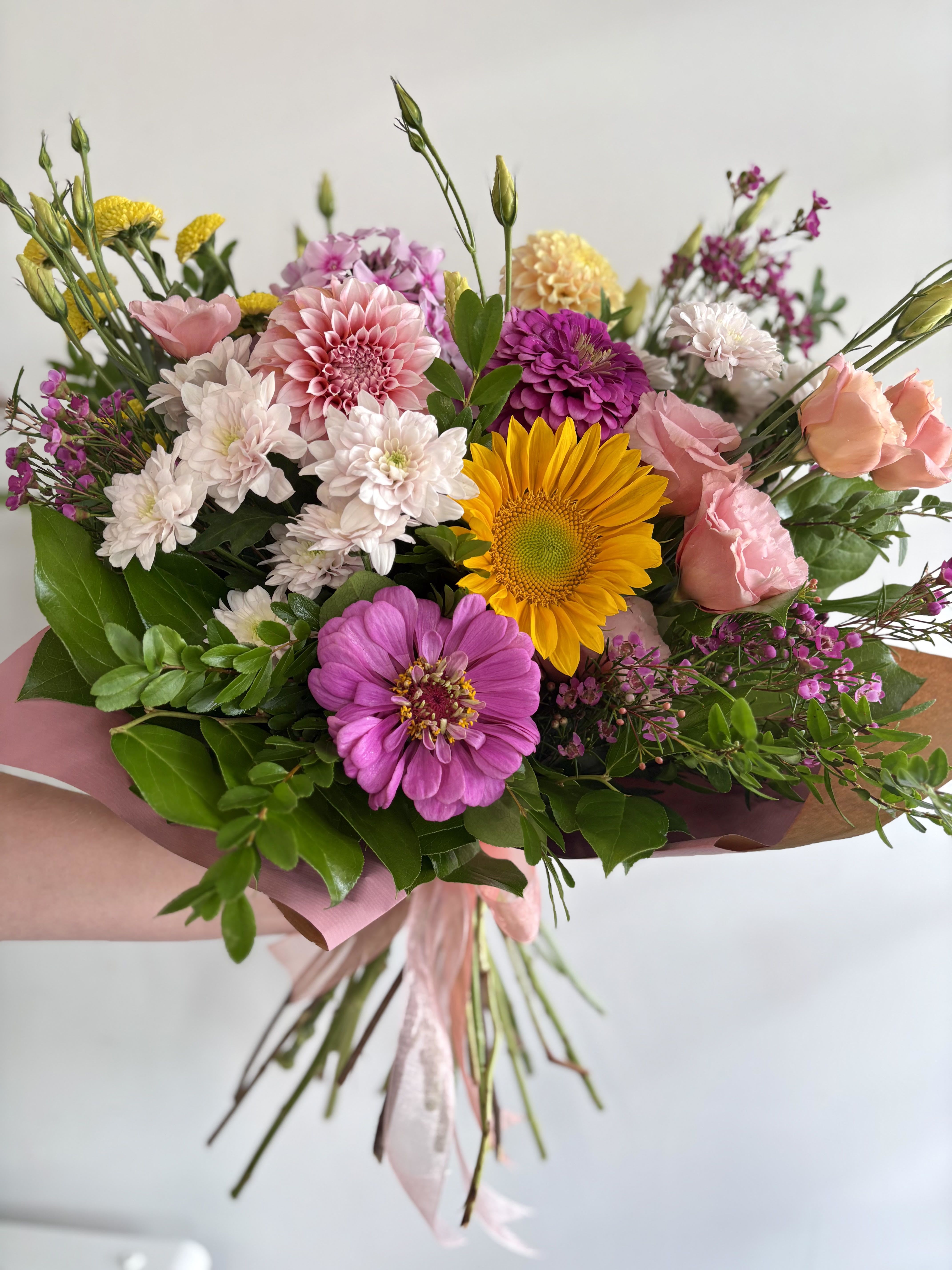 Bright hand-tied bouquet with pink roses, yellow sunflower, and mixed pink and white blooms wrapped in pink paper