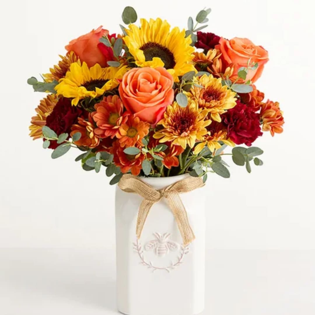 Bouquet of sunflowers, roses, and mums in a white vase with a ribbon