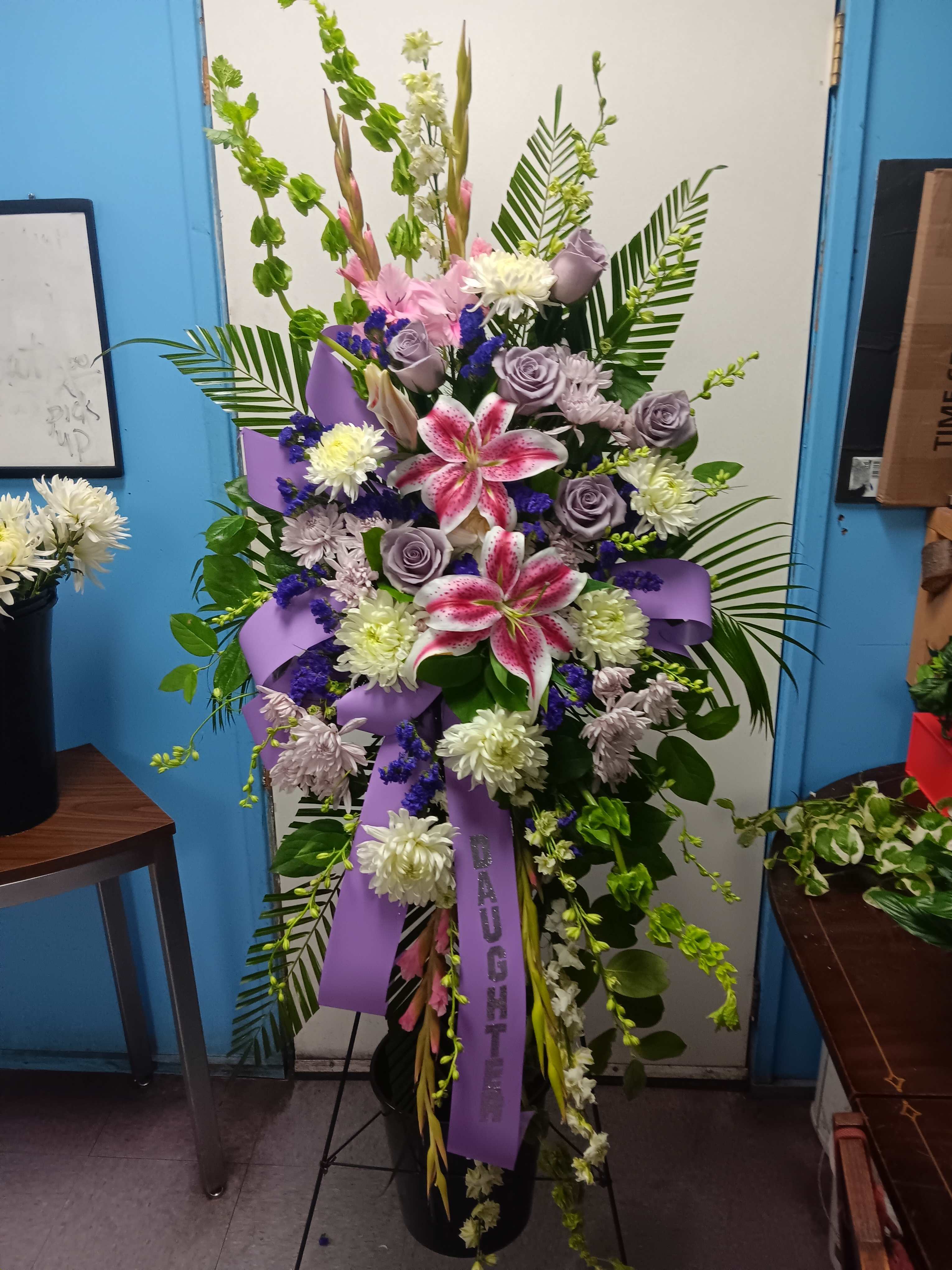 Tall floral spray with pink lilies, lavender roses, and purple ribbon in a black vase