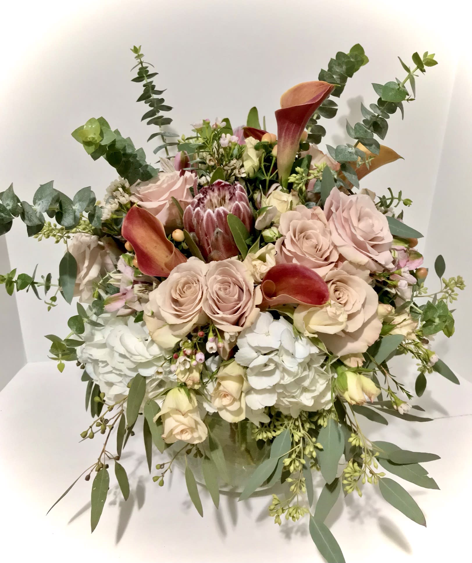 Round arrangement of blush roses, white hydrangeas, and orange calla lilies in a clear glass vase