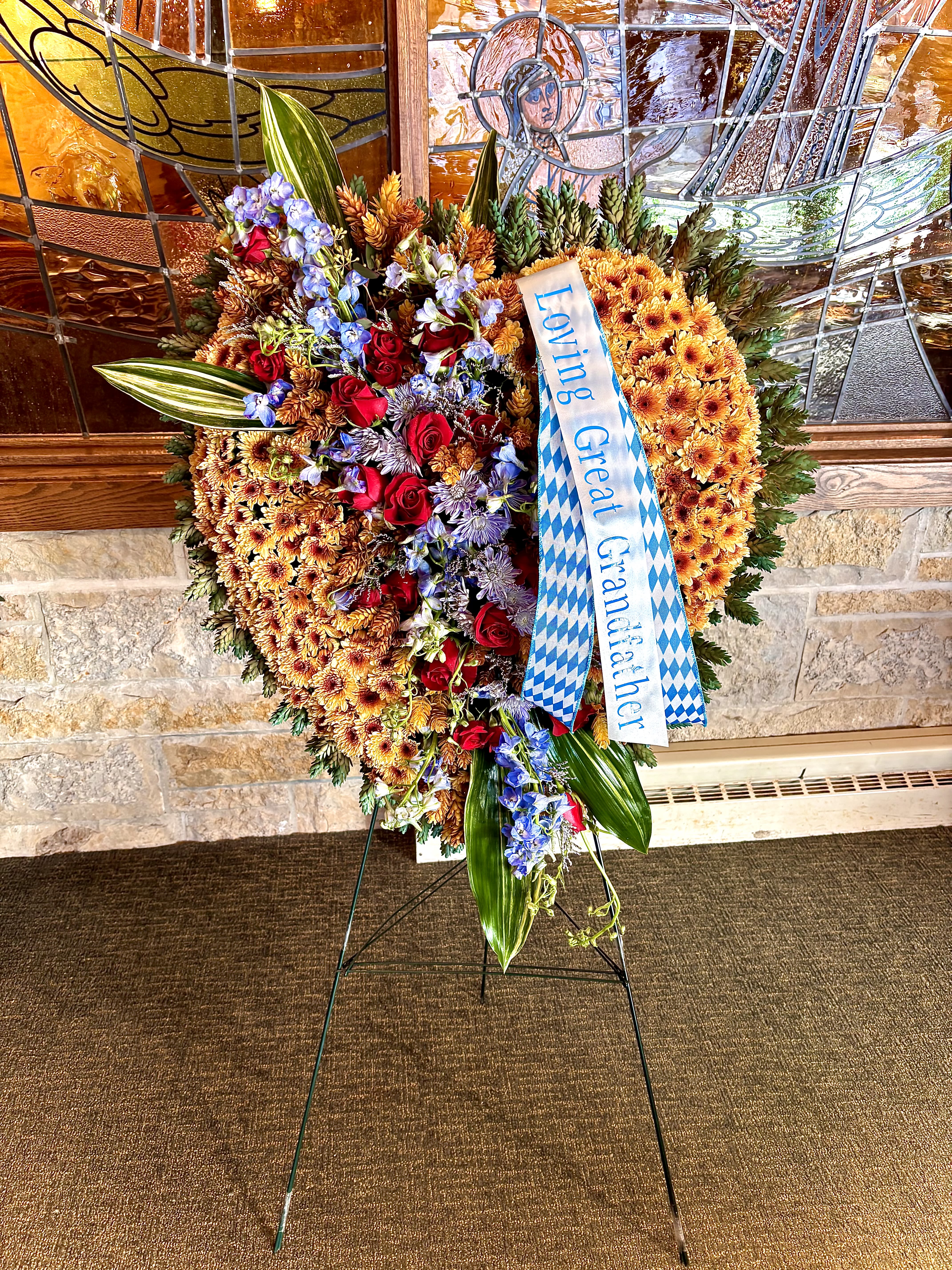 Heart-shaped standing funeral spray of orange mums with red roses, blue flowers, ribbon, and large green leaves on an easel