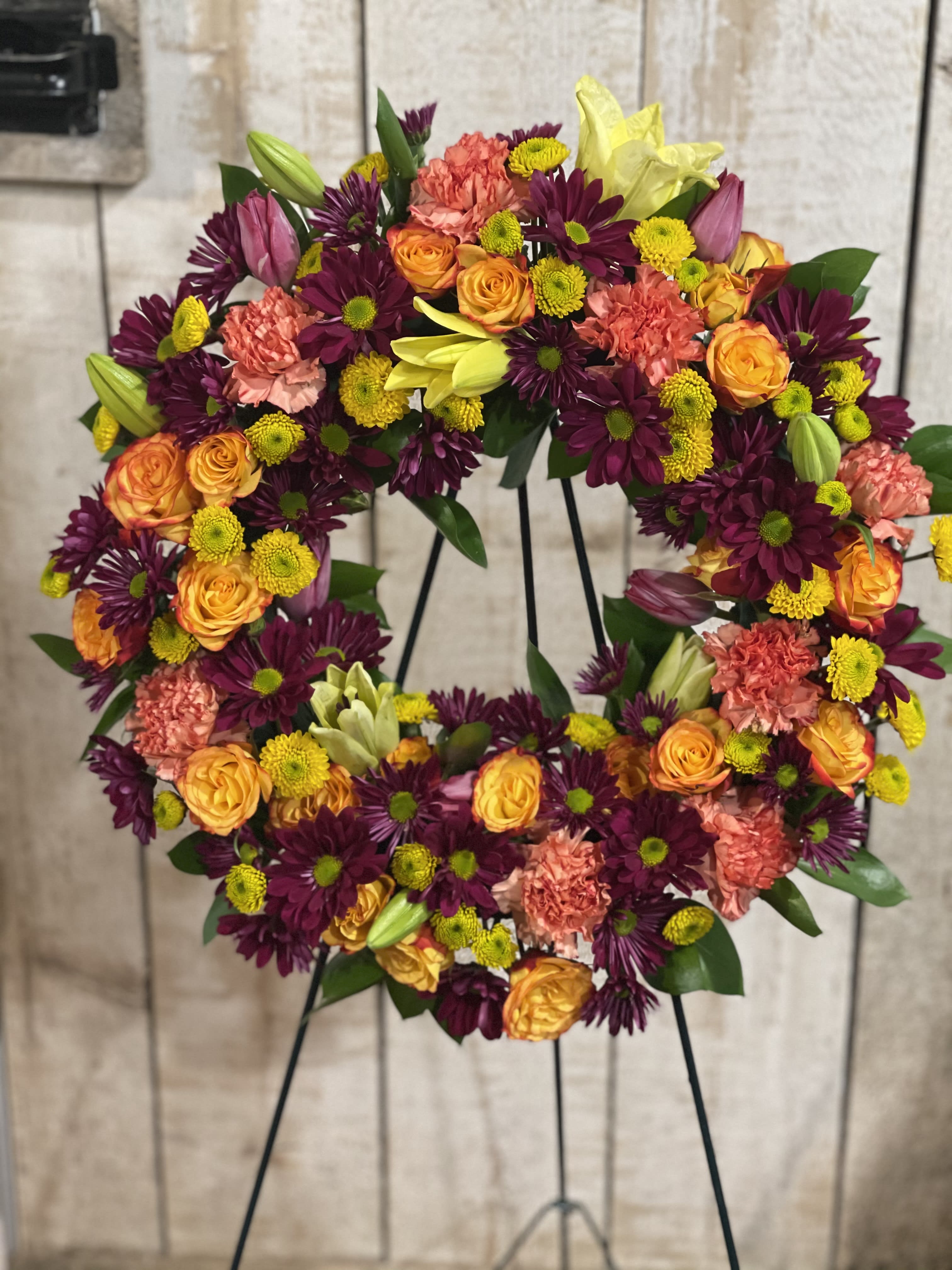 Floral wreath on a stand with orange roses, purple daisies, and pink carnations