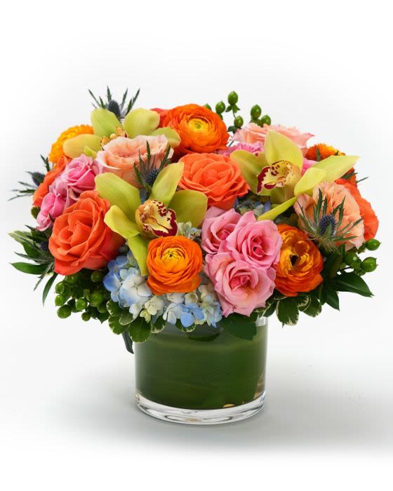 Low round arrangement of orange and pink roses, orchids, and ranunculus in a clear glass vase