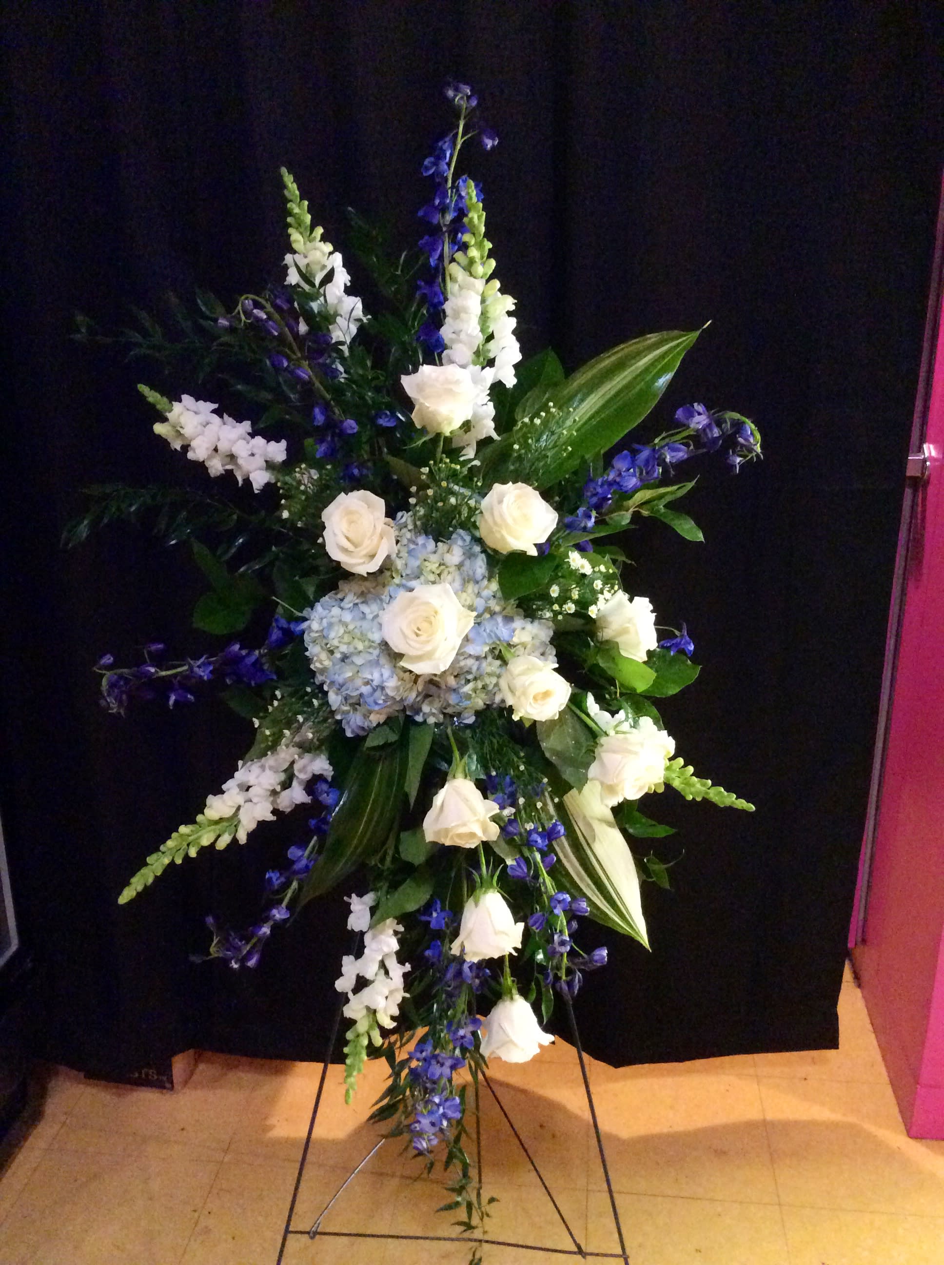 Tall standing floral spray with white roses and blue flowers on a black backdrop