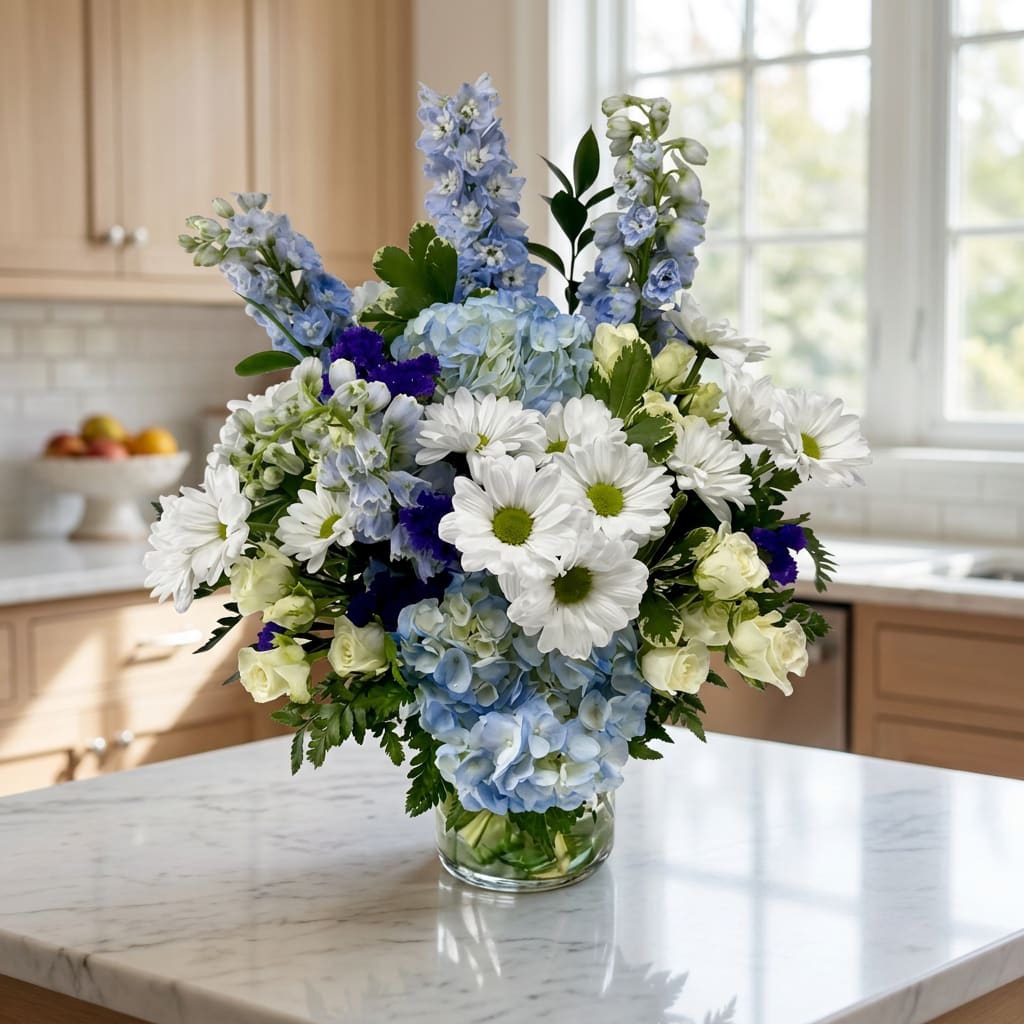 Sky Blue (DB) - Immerse yourself in a tranquil oasis with our enchanting Sky-Blue flower arrangement, showcasing a harmonious blend of blue hydrangeas, white spray roses, blue delphiniums, purple statice, white daisies and assorted greens. Artfully arranged in a leaf-lined glass cylinder vase, this stunning ensemble evokes a sense of serenity and beauty.