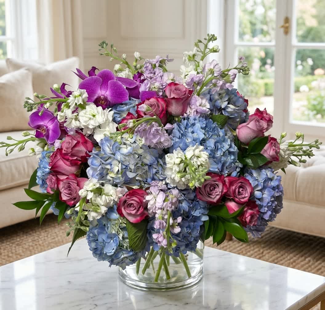 Grand Prize (DB) - Presenting an opulent display designed to make a lasting impression. This luxurious arrangement features a sophisticated blend of blue hydrangea, lavender roses, purple and white stock, lush greens, and filler flowers, all artistically arranged in an 8x8 glass vase.