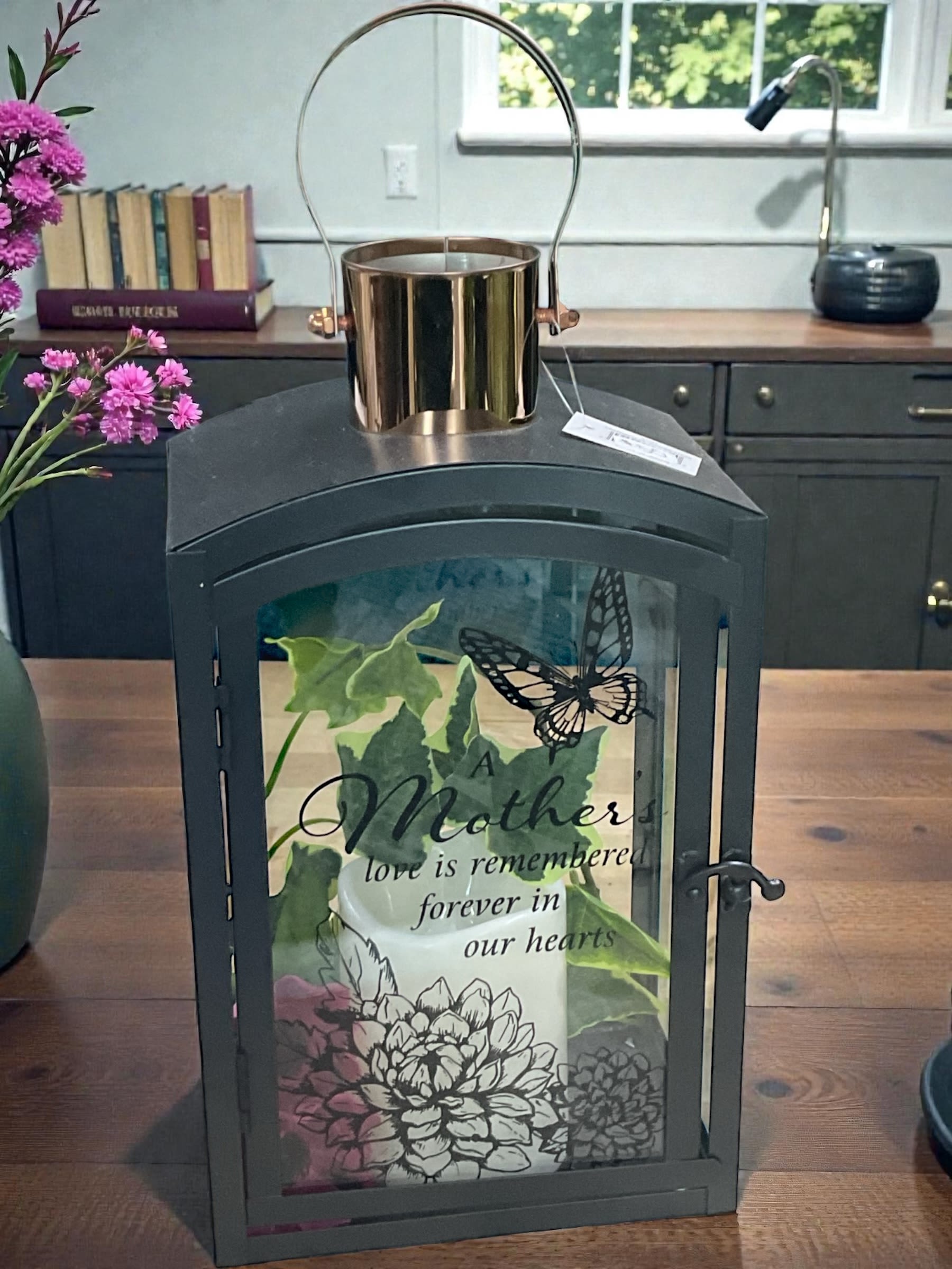 A Mother’s Love Lantern - Honor the memory of a beloved mother with our “A Mother’s Love is Remembered Forever in Our Hearts” memorial lantern. Featuring soft botanical artwork, delicate butterfly accents, and a heartfelt sentiment, this elegant keepsake offers comfort, remembrance, and a lasting tribute to a life filled with love.  Designed to provide a gentle glow and peaceful presence, this lantern is beautiful displayed in the home, on a covered porch, in a remembrance space, or as part of a memorial display. The classic black finish and timeless design make it a meaningful keepsake that can be enjoyed year after year.  The Standard version includes the lantern accented with a coordinating ribbon for a simple yet elegant presentation. Deluxe and Premium versions pair the lantern with fresh flowers selected to complement the lantern’s colors and sentiment. Premium designs feature additional flowers and enhanced texture for greater visual impact while maintaining a soft, graceful appearance.  A thoughtful choice for sympathy, remembrance, Mother’s Day, anniversaries of loss, celebrations of life, memorial gatherings, or simply honoring the memory of a cherished mother. This lantern offers a beautiful way to celebrate a life, cherish treasured memories, and keep a mother’s love close to the heart for years to come.