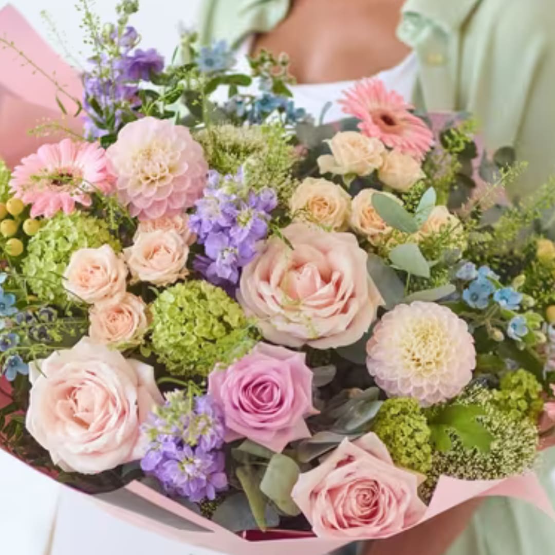 Pastel bouquet of roses, dahlias, and purple blooms wrapped in pink paper