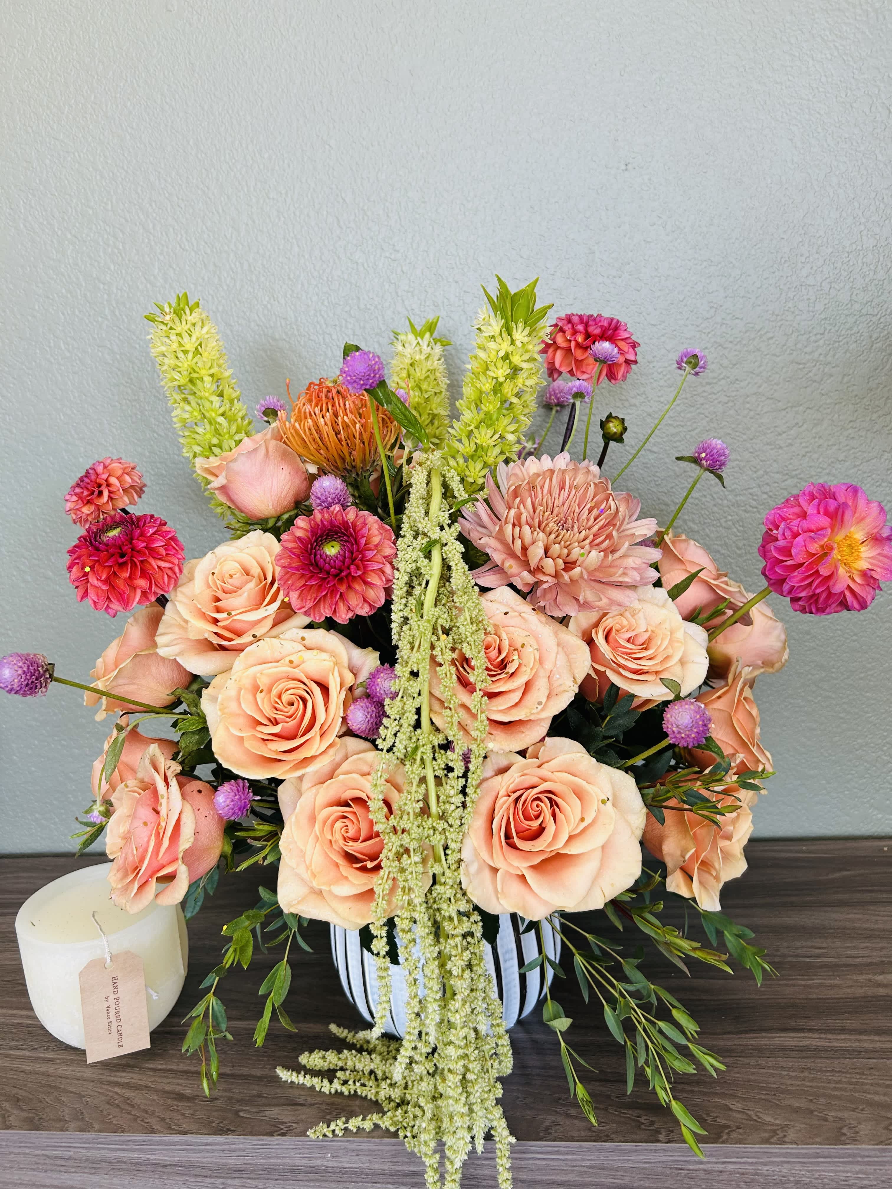 Peach roses and pink blooms arranged in a striped vase with a candle beside it