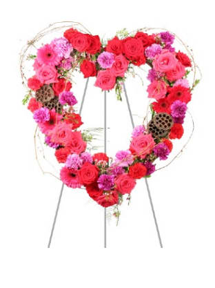 Heartfelt Array Standing Spray - A bold heart formed from tightly clustered pink, red, and purple blooms makes an immediate statement-the perfect heart-shaped standing spray for a memorial service or a love-themed celebration. This flower arrangement and standing spray combines roses, gerbera-style daisy blooms, and carnations into a dense, velvety surface that reads romantic and respectful at once, so the person you're honoring will feel seen and remembered.  The composition sits on a sturdy metal easel stand and is accented with dried lotus pods and airy decorative twigs for a touch of rustic contrast against the fresh petals. Rose heads mingle with daisy-like gerberas and ruffled carnations to create rich texture and depth; the pinks and reds deliver warmth while the purples add a subtle, somber glow. Sized to read as a statement piece, it's designed to be viewed standing-ideal for ceremony aisles, memorial displays, or a dramatic tribute at a celebration of life.  Choose this standing spray when you want to send a clear message of love, sympathy, or celebration-the heart shape says it plainly for you. Care is simple: because it's arranged for upright display, keep it sheltered from direct sun and wind at the venue; if delivery timing is important, select the appropriate service to ensure it arrives fresh for the occasion.