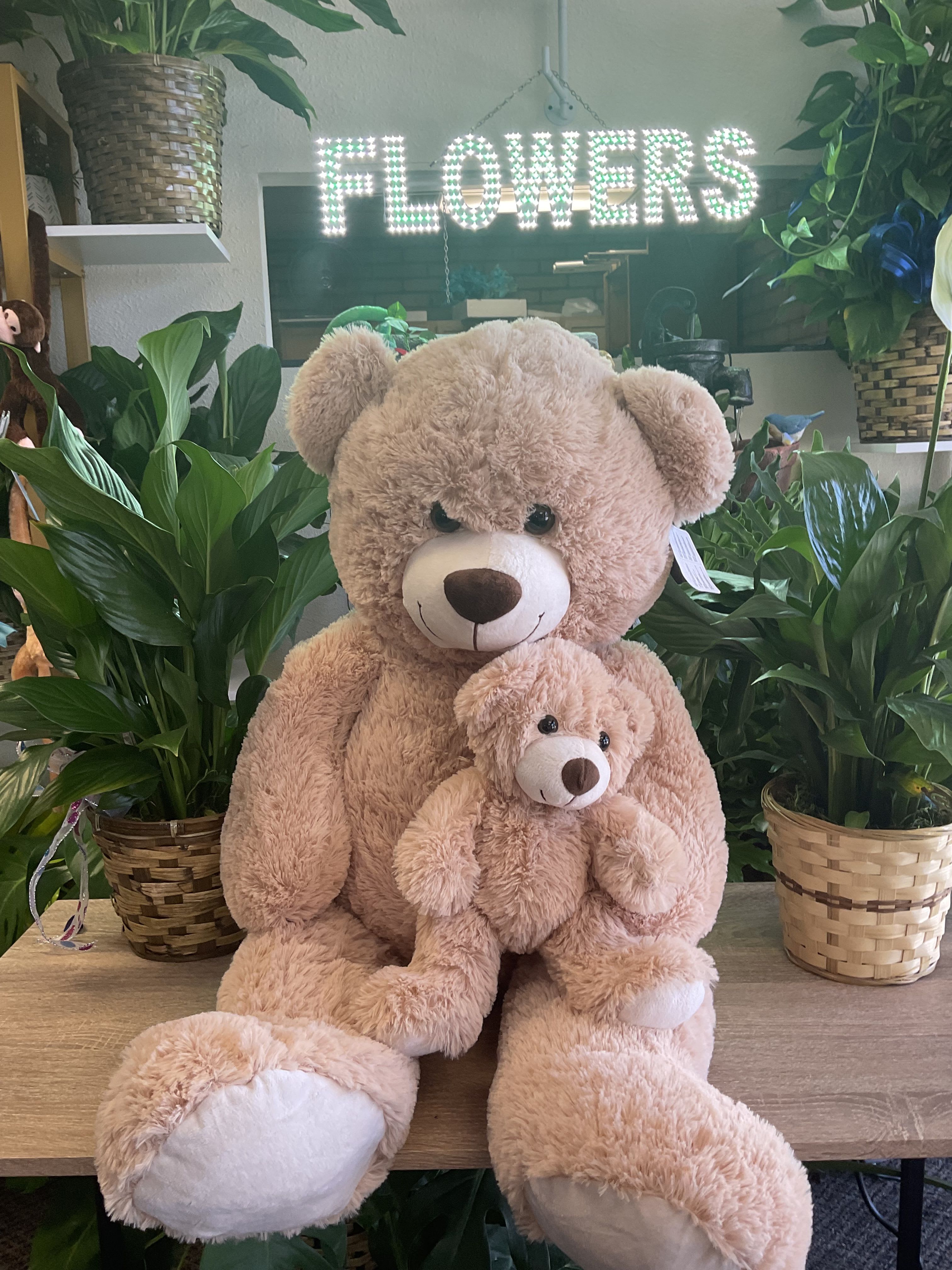 Two plush teddy bears sit among potted plants under a lit "FLOWERS" sign.