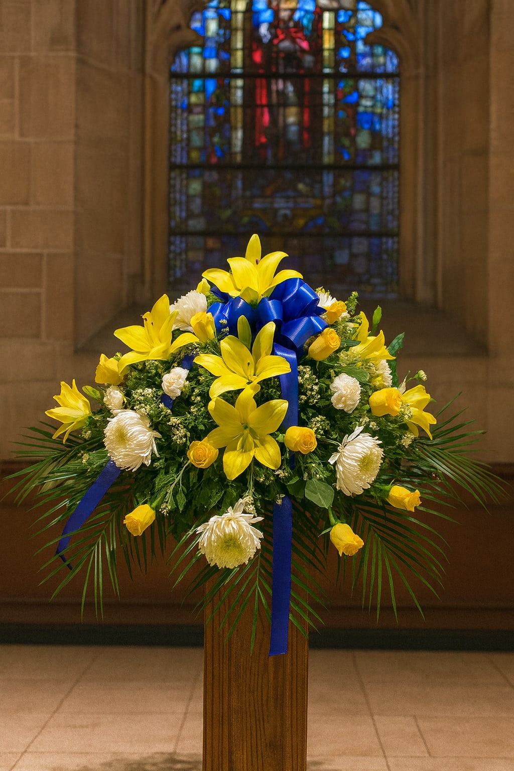Yellow lilies and white flowers arranged with a blue ribbon on a pedestal.