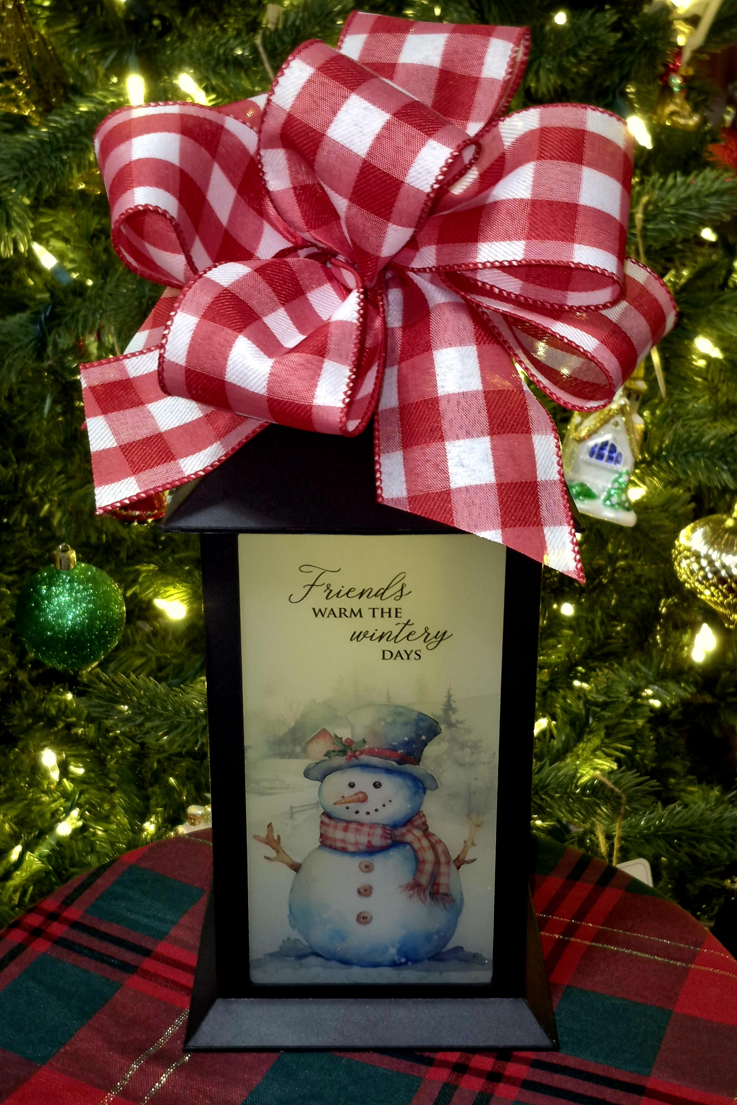 Gift box with a red plaid bow and a snowman card in front of a Christmas tree