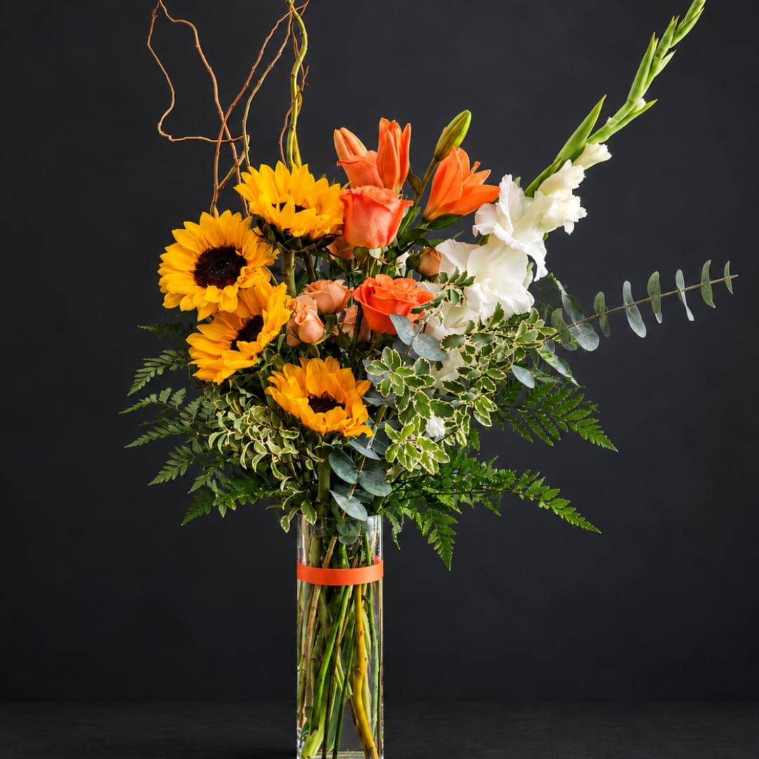 Tall bouquet of sunflowers, orange roses, and white gladiolus in a glass vase