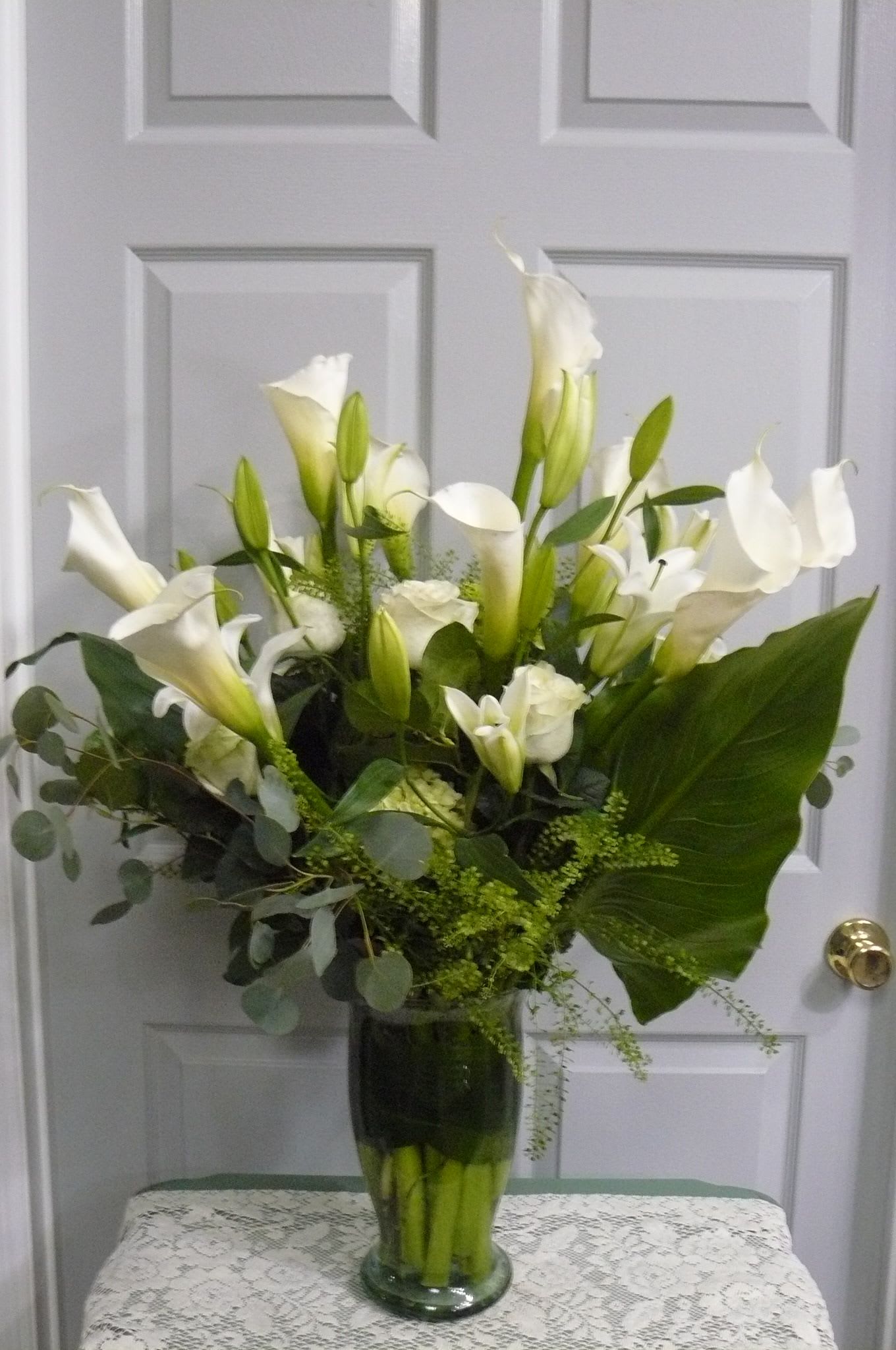 Tall arrangement of white calla lilies and roses with a large green leaf in a clear glass vase