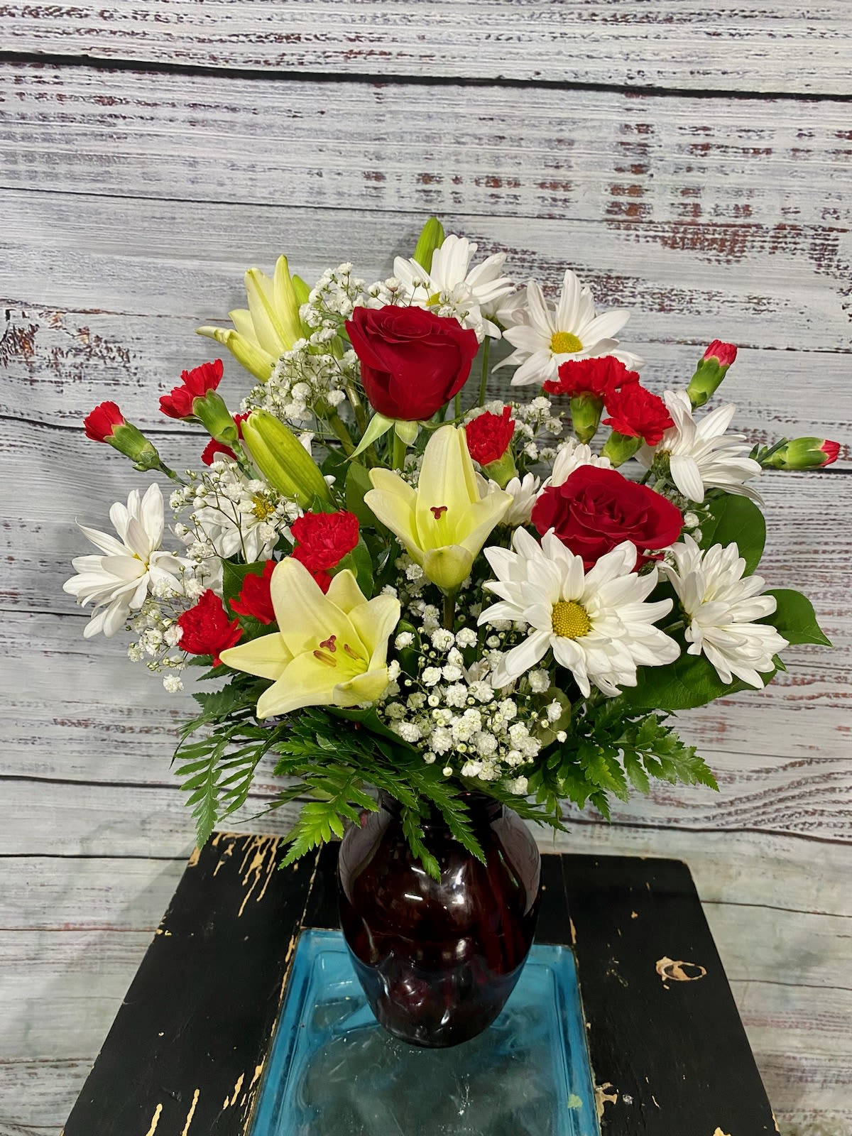 Be My Love Bouquet - A vibrant mix of red roses and red carnations paired with pale yellow Asiatic lilies and crisp white daisies creates a classic romance-forward arrangement ideal for an anniversary bouquet. The strong red tones send a clear message of love, while the soft whites and yellows temper the drama so the person you're thinking of will feel both celebrated and comforted - perfect for holiday gifting, a "just because" gesture, or any moment when you want to say I love you.  This rounded arrangement is designed to sit upright and full in a dark glass vase that gives the composition visual weight and a polished, modern look. Airy baby's breath and fresh greenery tuck between the focal blooms to add texture and lift; the overall silhouette is neat and generous rather than loose. Colors include vivid red, pale yellow, clean white, and fresh green to suit both romantic and everyday occasions.  Send this bouquet to mark anniversaries, congratulate someone special, or brighten Mother's Day with a confident, cheerful statement. It arrives ready to display in the included dark glass vase - trim stems and fresh water as needed to keep the lilies and roses looking their best.