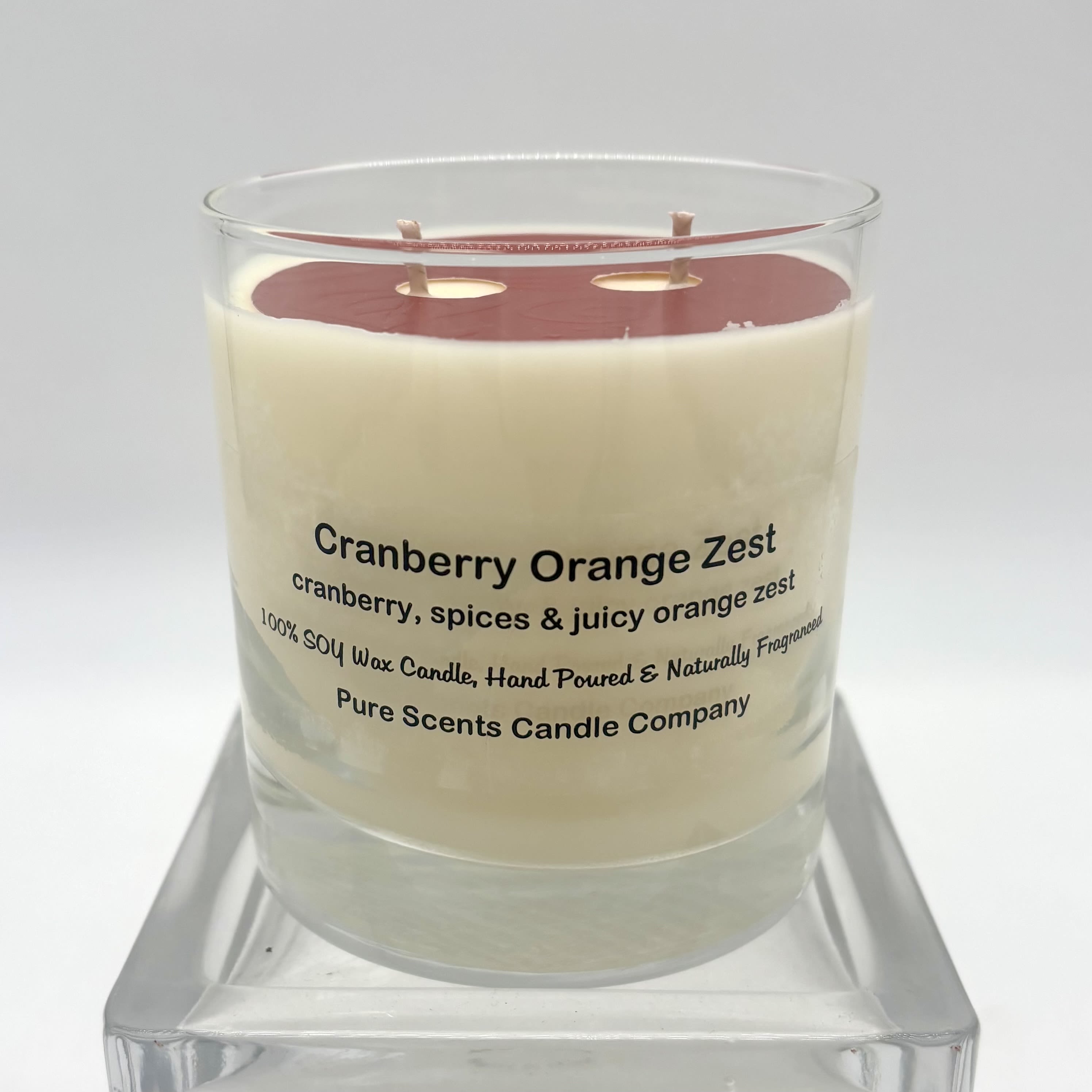 Glass jar candle labeled Cranberry Orange Zest with cream wax and red top layer on a clear stand