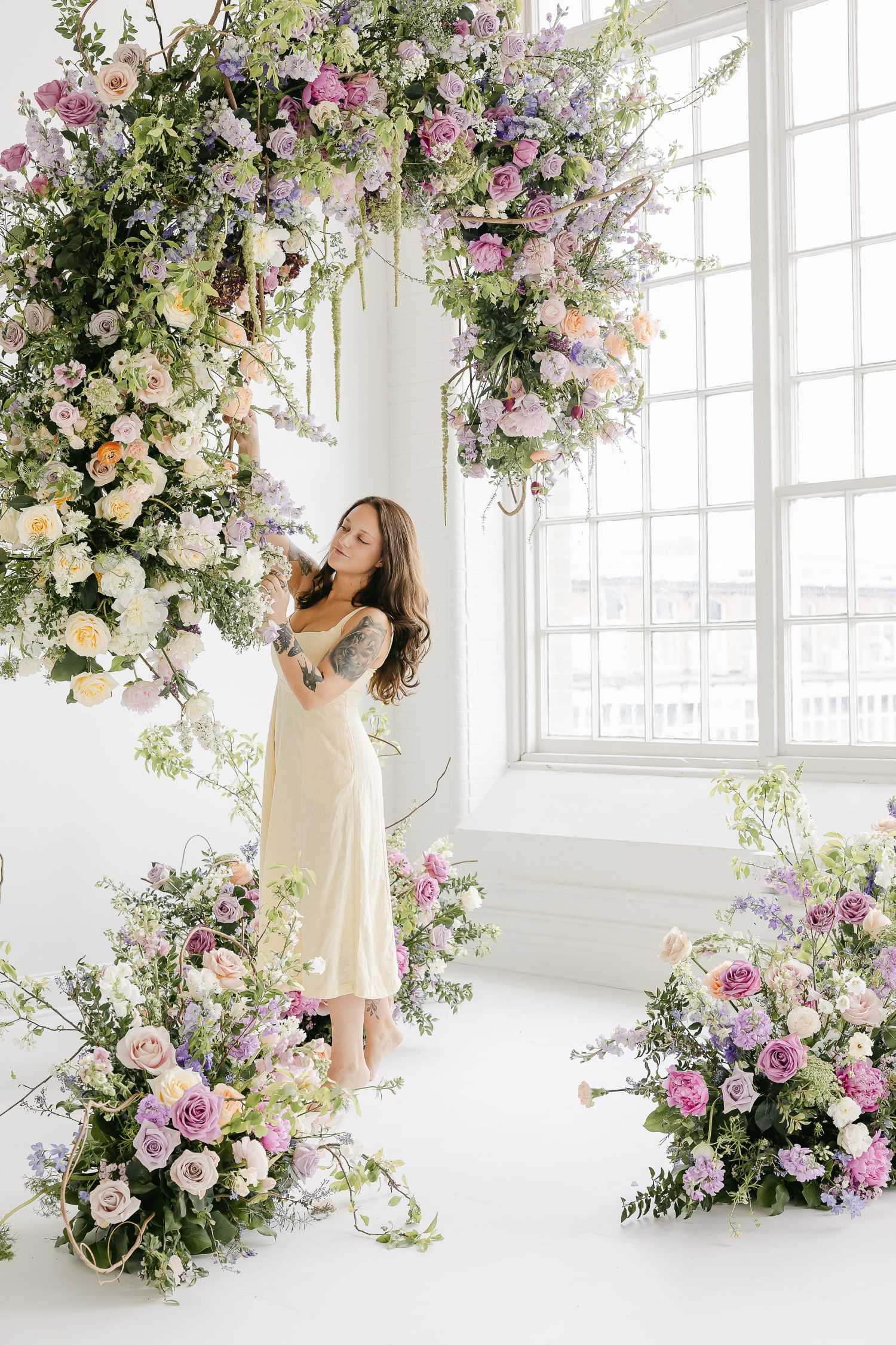 Kristina posed standing and reaching up to touch the flowers on a large hanging floral installation, surrounded by floral meadow arrangements on the floor.